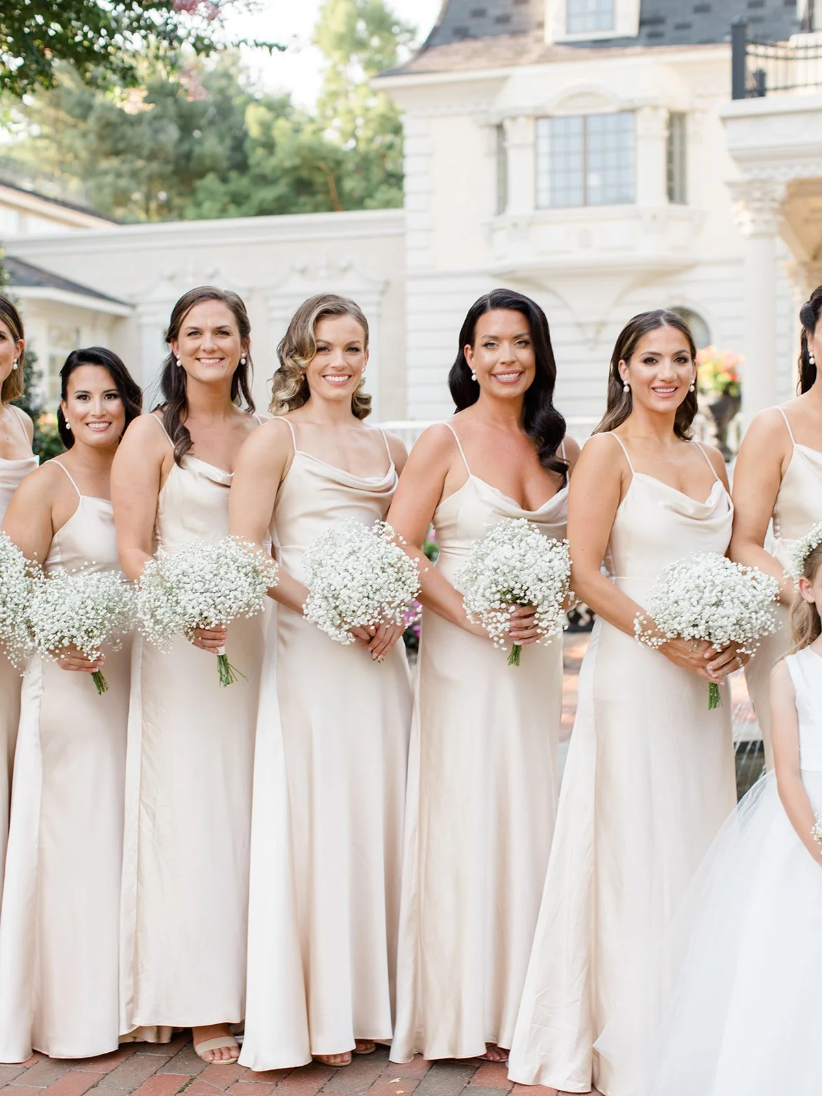 Bridesmaids in champagne-colored dresses holding white flower bouquets at a wedding.