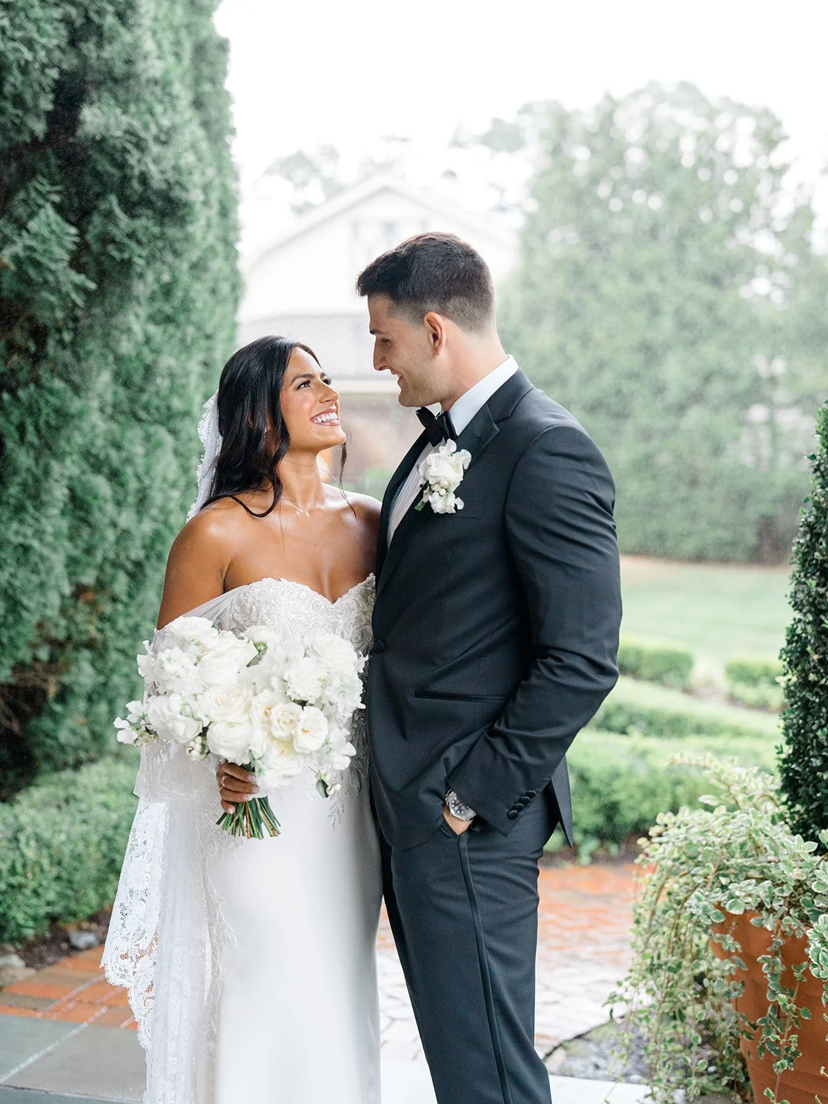 A bride and groom looking at each other and smiling at each other outside. The bride is holding a bouquet and dressed in a white wedding gown. The groom is in a black tuxedo with a bow tie. They are standing on a pathway surrounded by greenery.