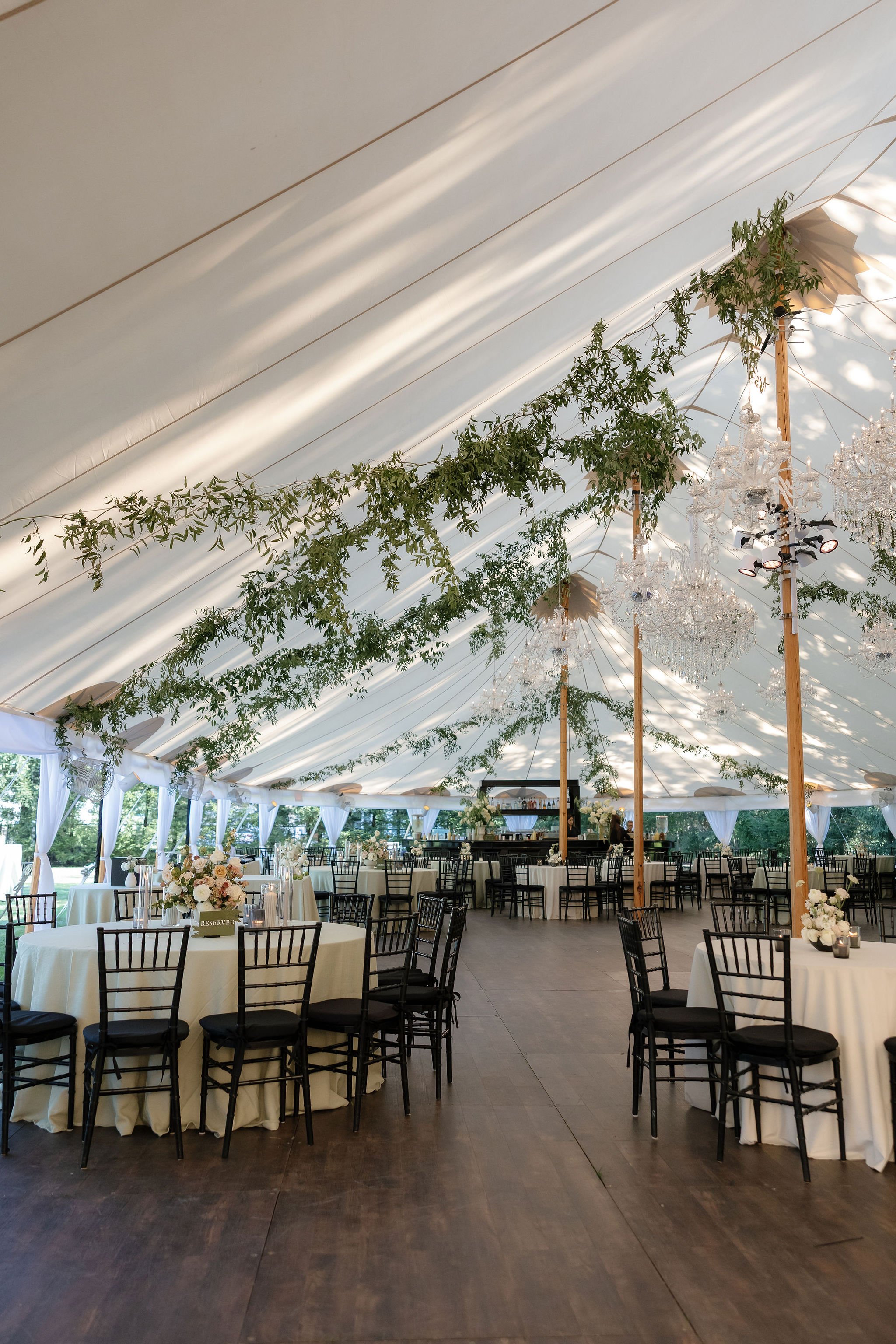 An elegantly decorated event tent with round tables draped in white tablecloths, black chairs, and floral centerpieces. Greenery and chandeliers hang from the ceiling, creating a festive atmosphere.