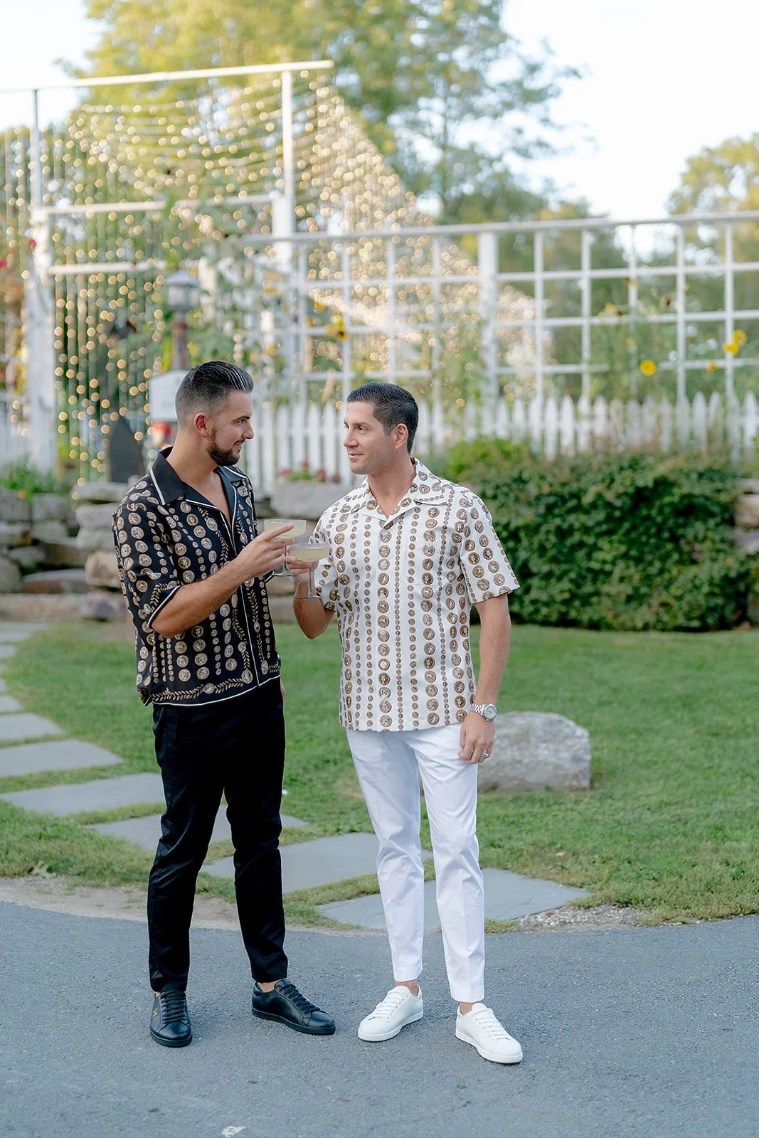 Two men in patterned shirts holding drinks and talking outdoors with string lights and greenery in the background.