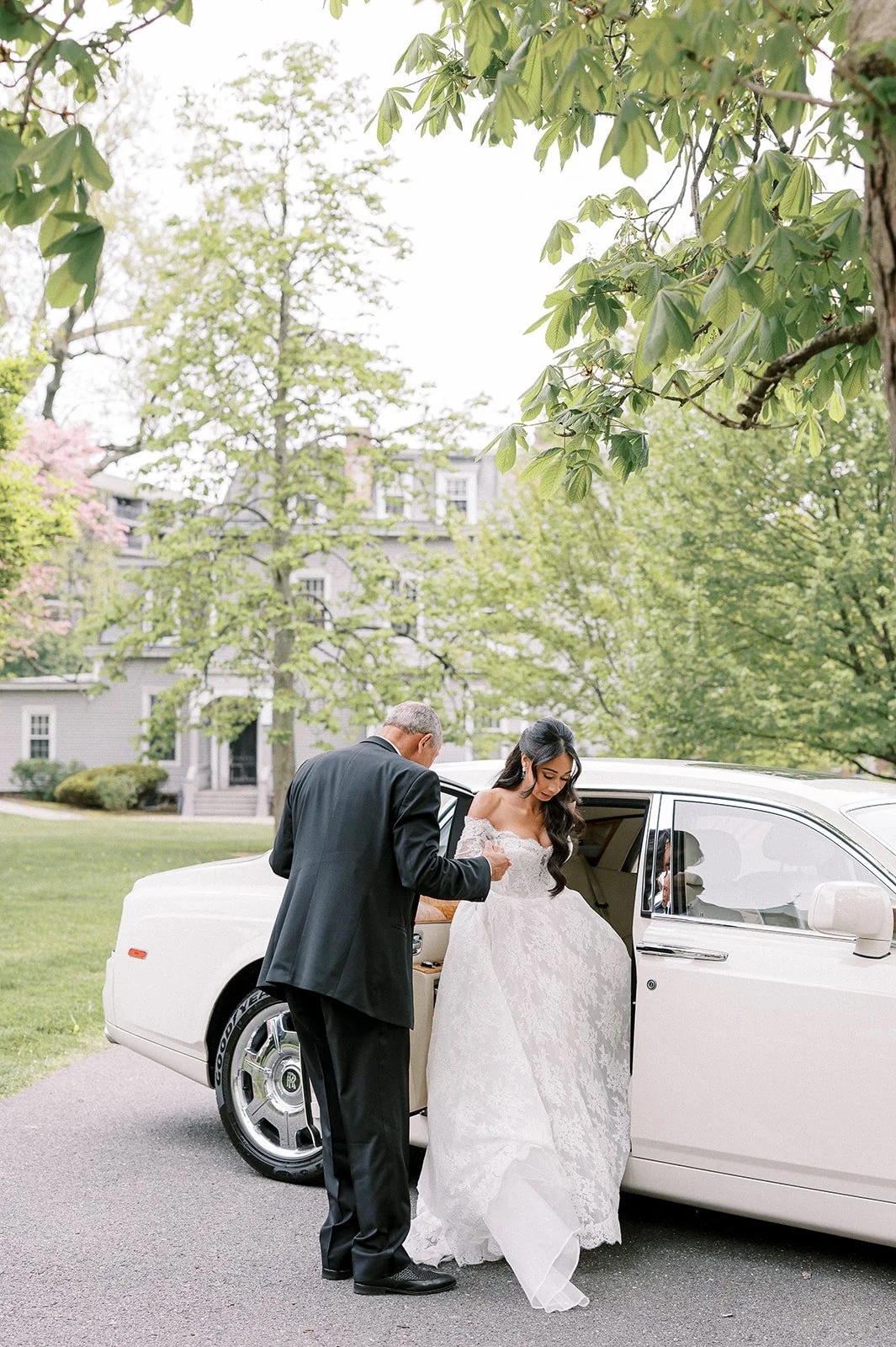 A bride in a white wedding dress and an older man in a black suit standing outside next to a white limousine.