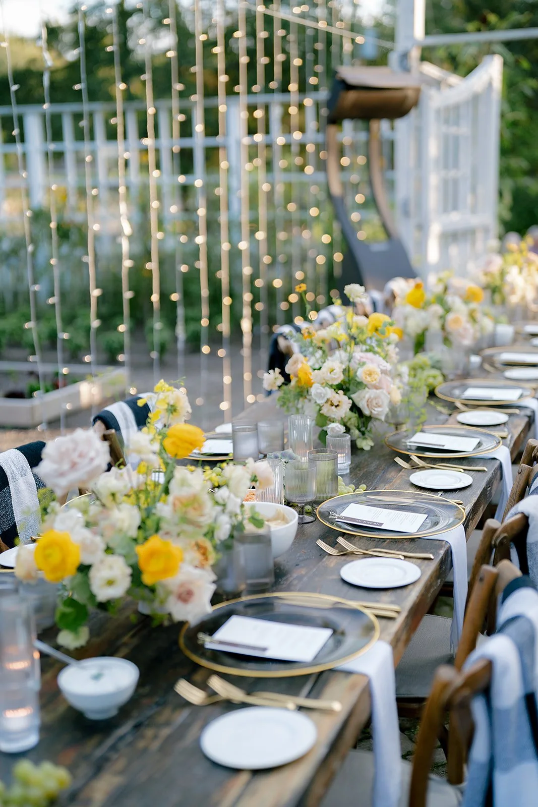 Outdoor dining table decorated with white and yellow flowers, candles, and elegant tableware, set against a backdrop of string lights and greenery.