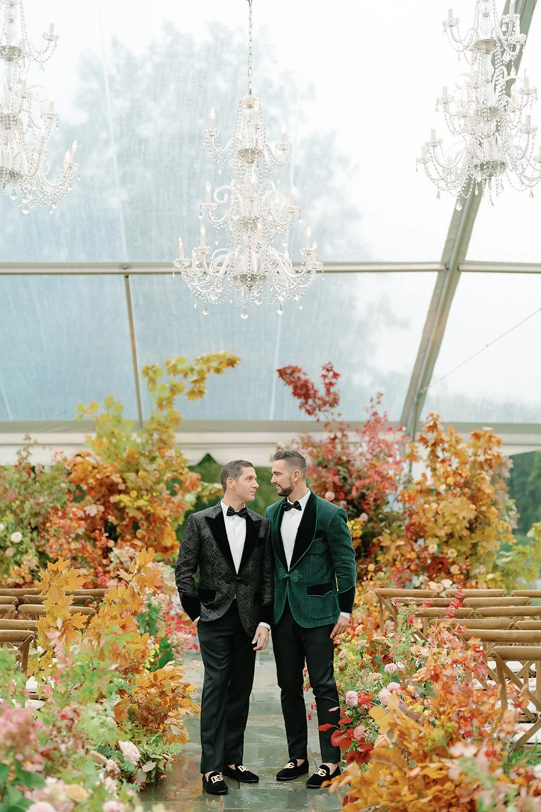 Two men in tuxedos standing close together in a decorated reception venue with colorful autumn foliage and chandeliers overhead.