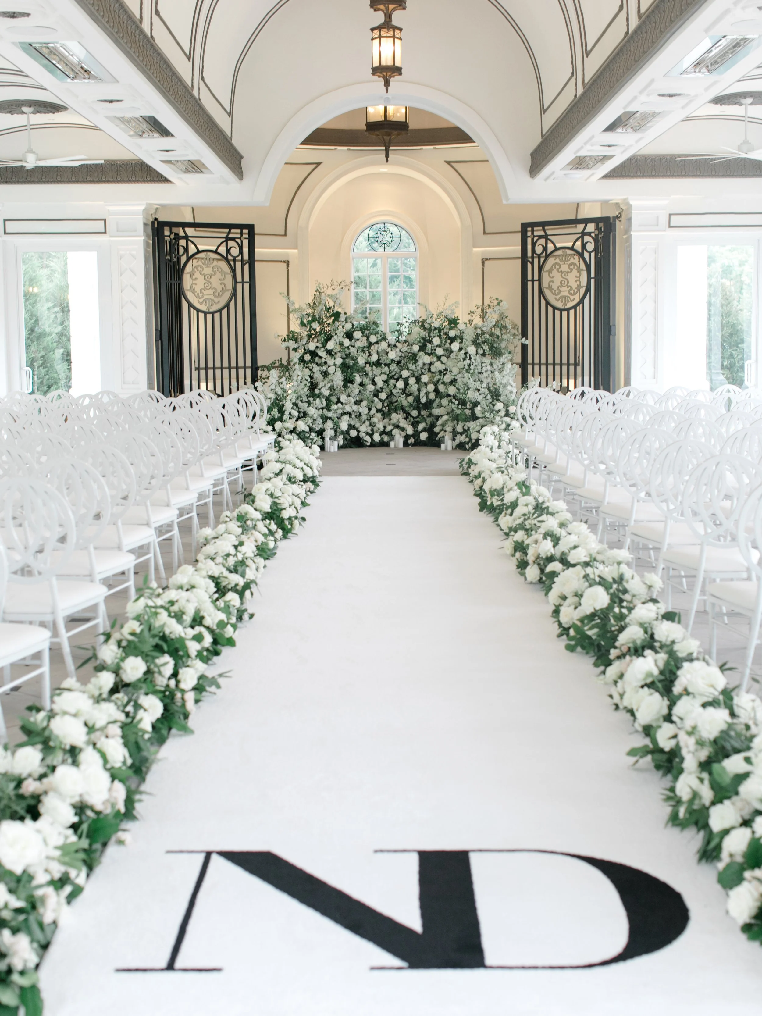 A wedding aisle decorated with white flowers on both sides, leading to a floral backdrop near a window in an elegant room.