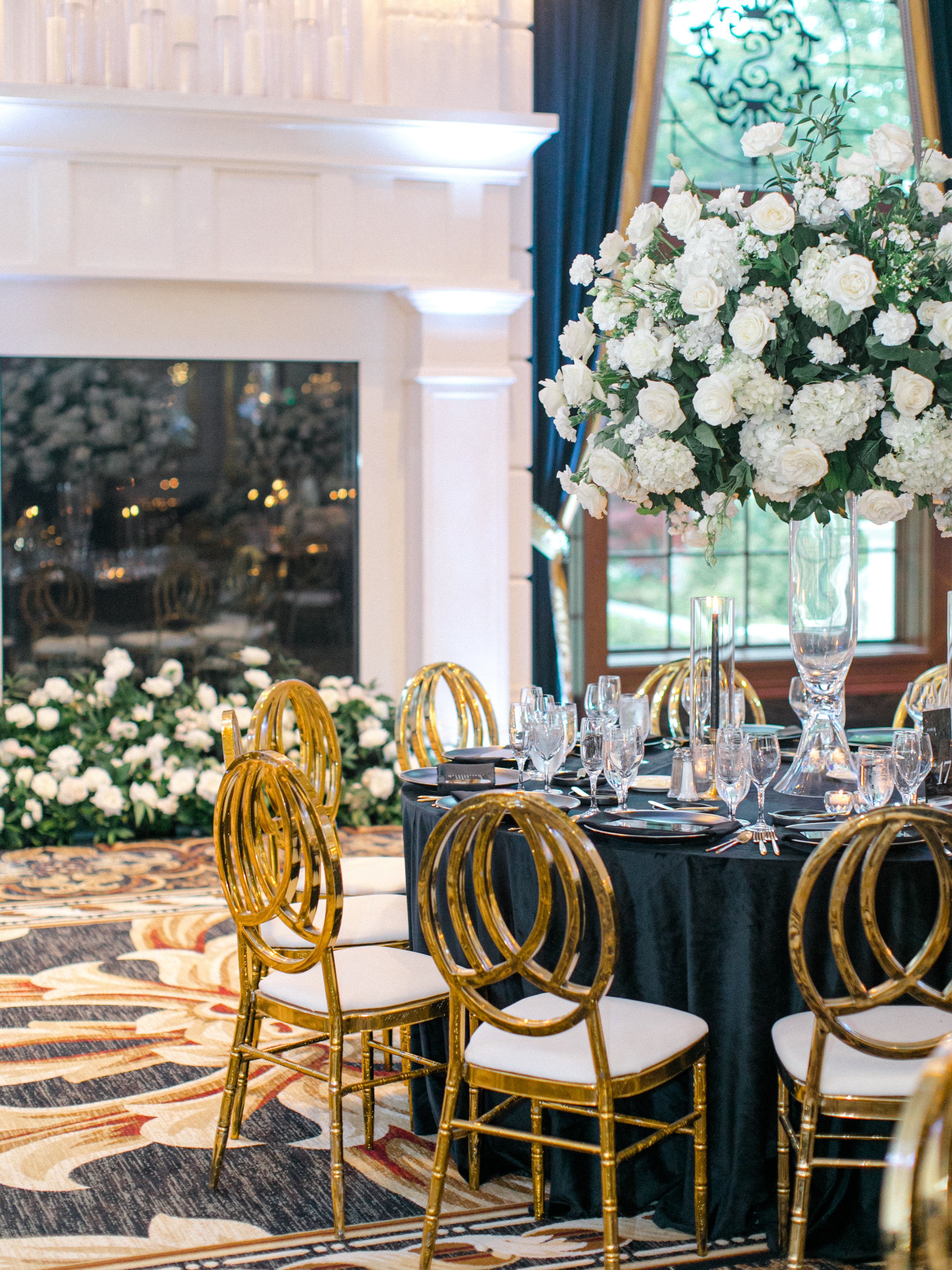 Elegant banquet table setup with gold chairs, dark tablecloth, fine glassware, and large floral centerpiece in a room with a white fireplace, large windows, and dark curtains.