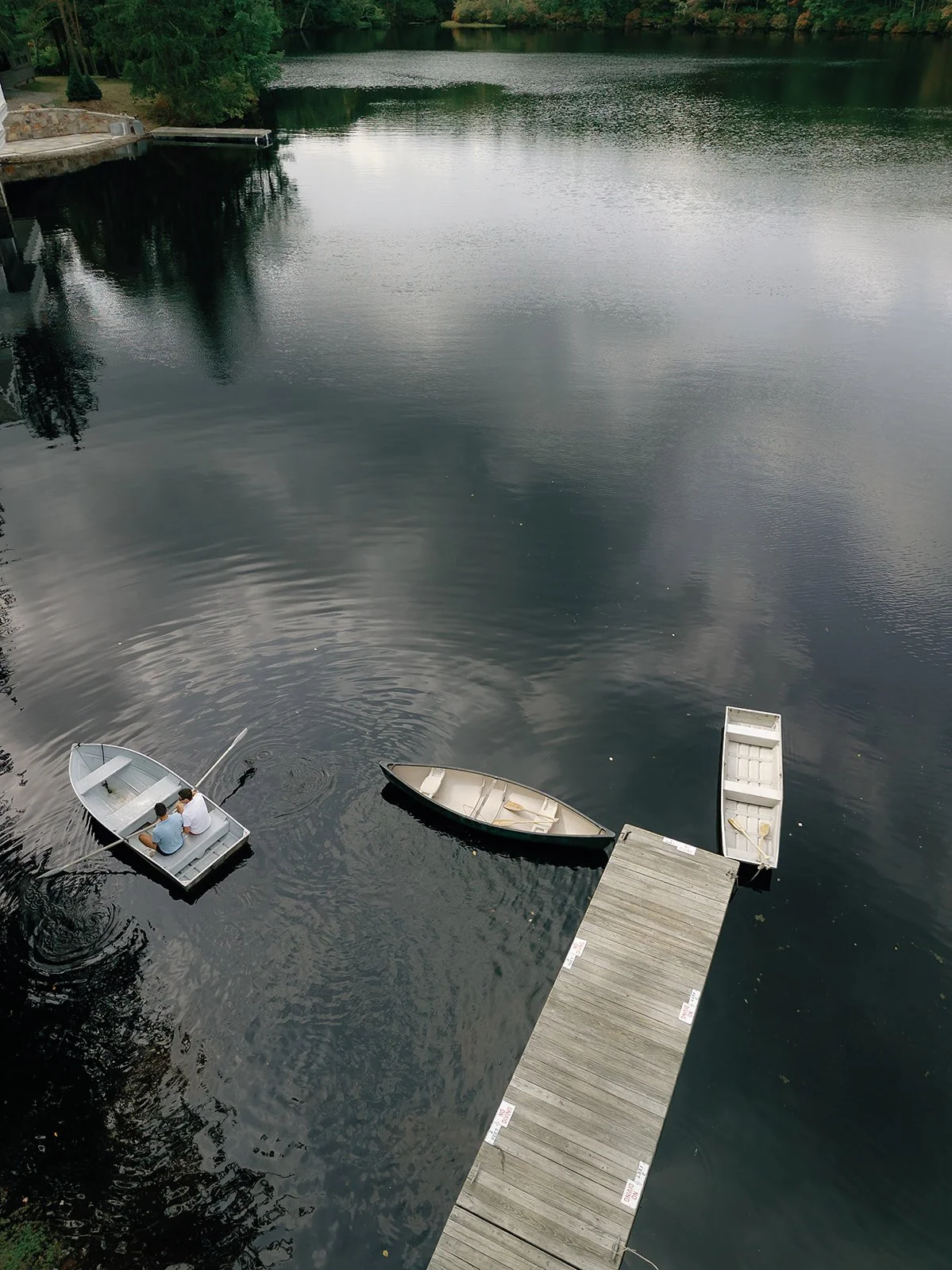 Two people sitting in a small rowboat on a calm river near a wooden dock, with two empty rowboats tied to the dock, surrounded by trees and overcast sky reflected in the water.