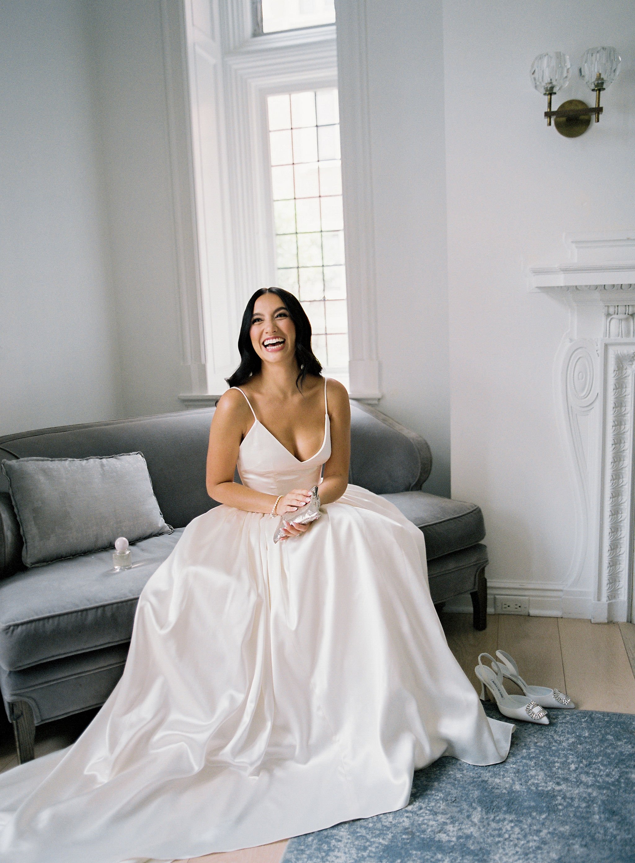 A woman in a white dress sitting on a gray sofa, smiling and laughing, in a bright room with large windows, white walls, and a fireplace. She has black hair, and white high-heeled shoes are on the floor beside her.