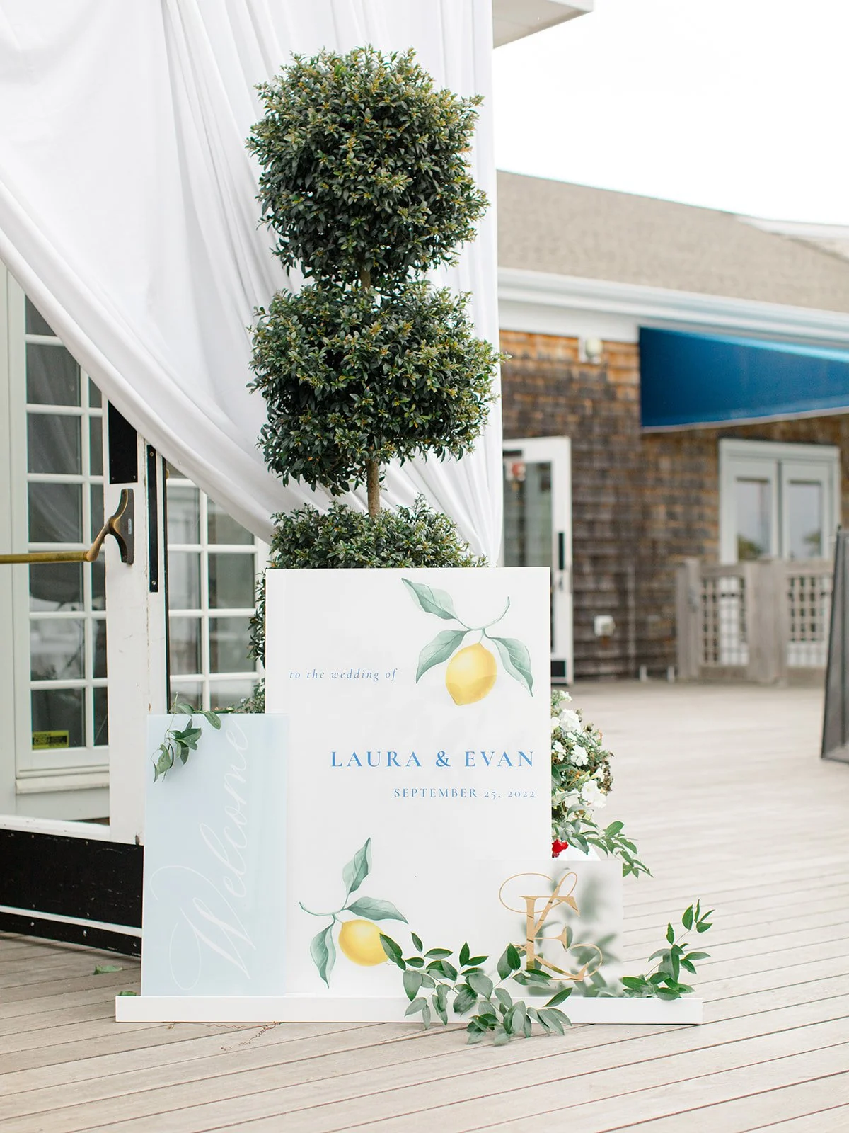 Wedding sign with lemon illustrations, announcing the wedding of Laura and Evan on September 25, 2022, decorated with greenery and flowers, positioned on a wooden deck near a white curtain and potted topiary
