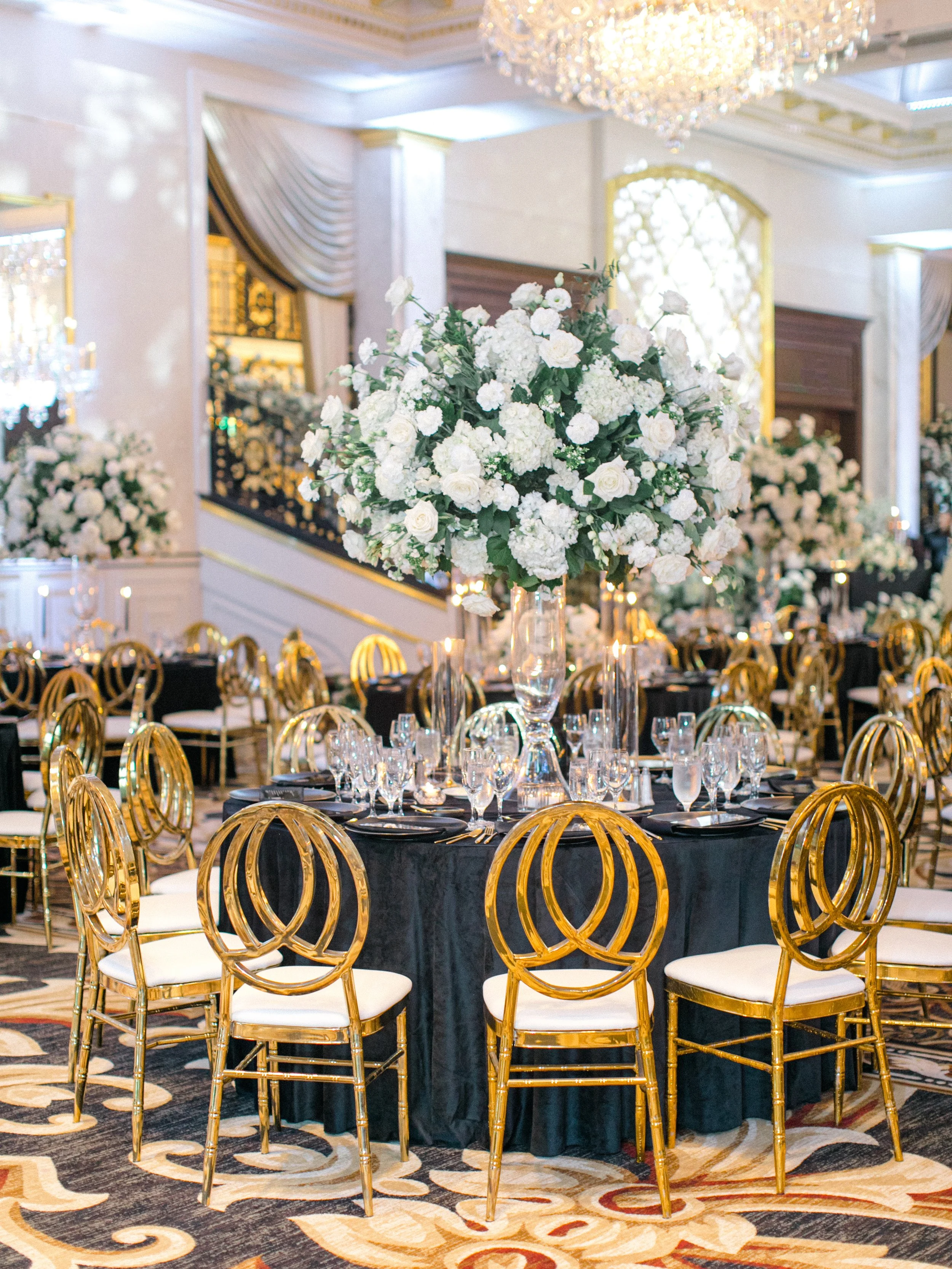 Elegant banquet table setup with a large floral centerpiece, gold chairs, black tablecloth, fine glassware, and candelabras in a luxurious decorated room with chandelier.