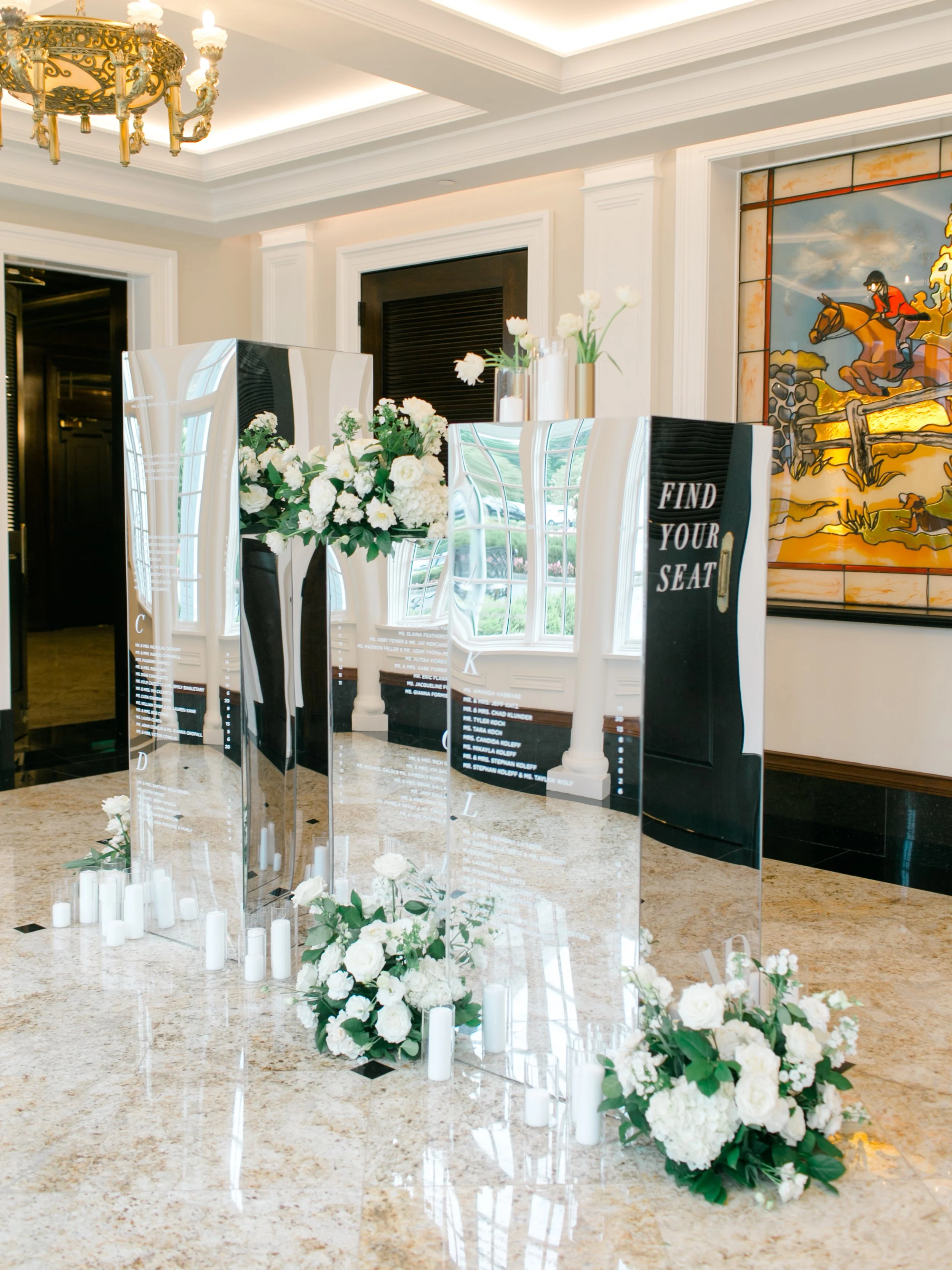 Mirror with white floral arrangements and candles on a marble floor, with a sign saying 'Find Your Seat' in a decorated hotel lobby or event space.