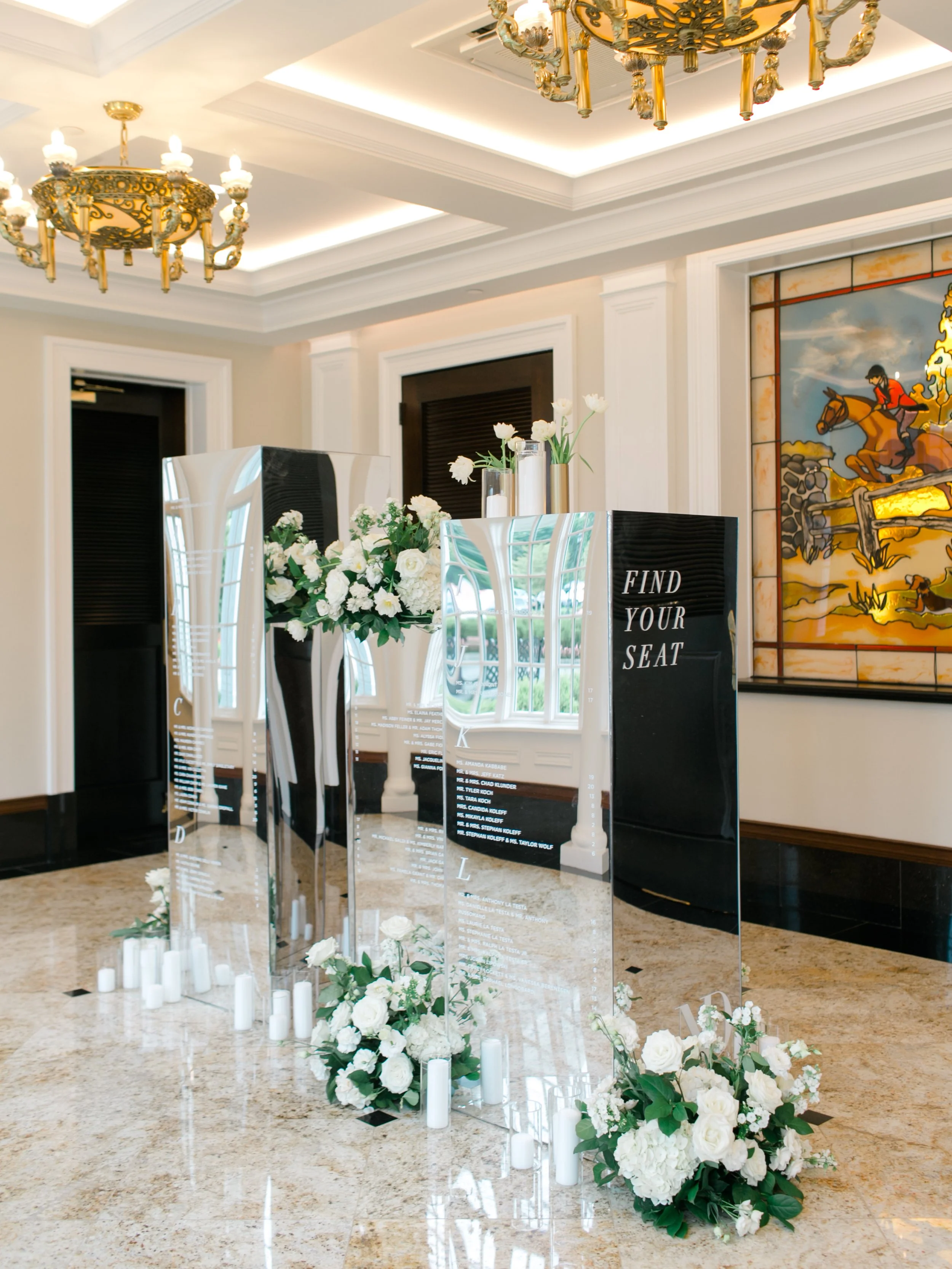 Elegant wedding or event display with three mirrored boards, white flower arrangements, and candles, set in a decorated room with ornate chandeliers and a stained glass window.