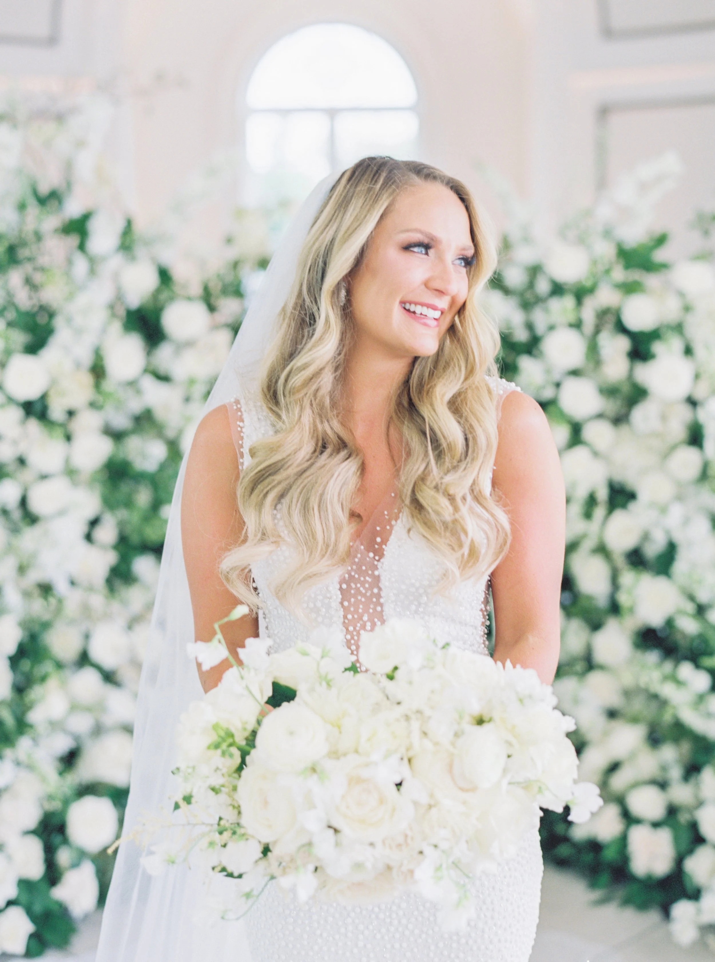 A bride in a white wedding dress holding a bouquet of white flowers, smiling inside a decorated venue with white flowers in the background.