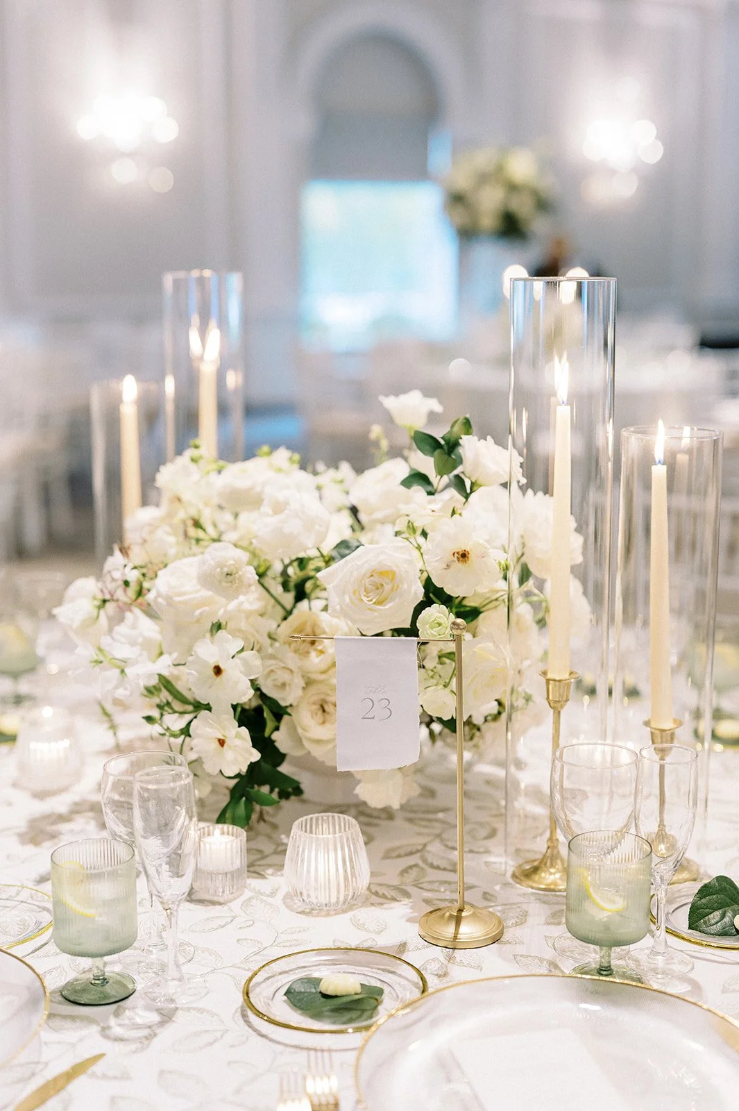 Elegant wedding table decor with a large white floral centerpiece, tall glass candle holders with lit candles, small votive candles, and glassware on a white embroidered tablecloth.