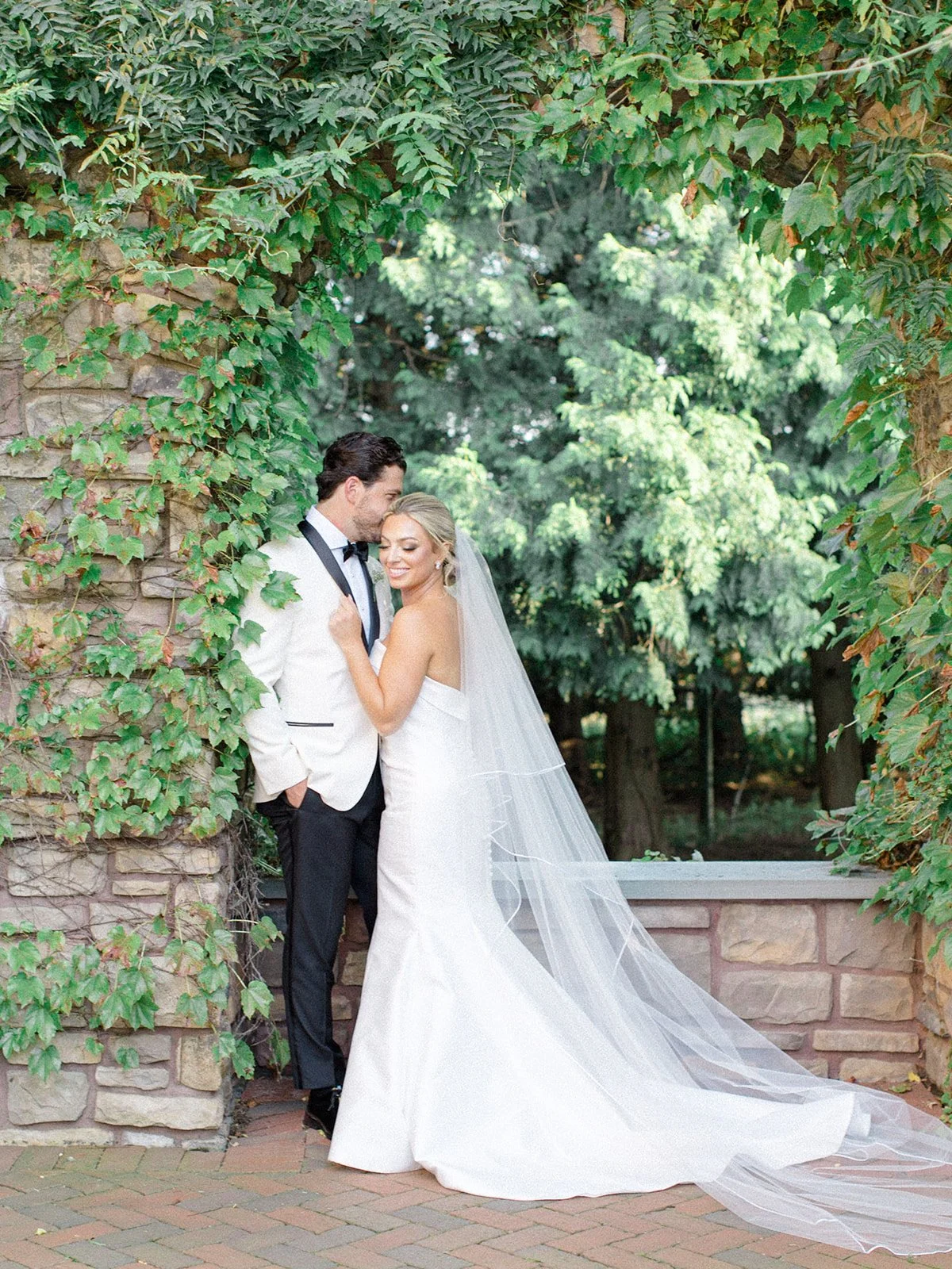 A bride and groom stand close together under green foliage, smiling. The bride wears a white wedding dress with a long veil, and the groom wears a white tuxedo jacket with black pants and a black bow tie. They appear happy in a romantic outdoor setti