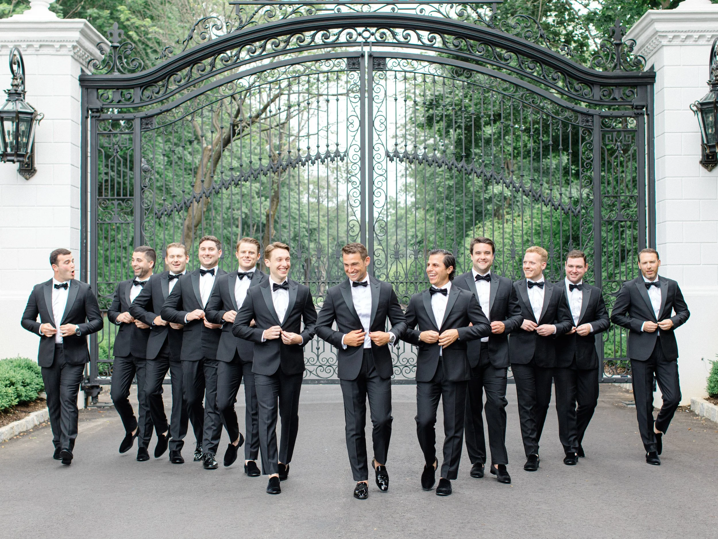 A group of eleven men in tuxedos walking together in front of a large decorative metal gate, smiling and laughing.