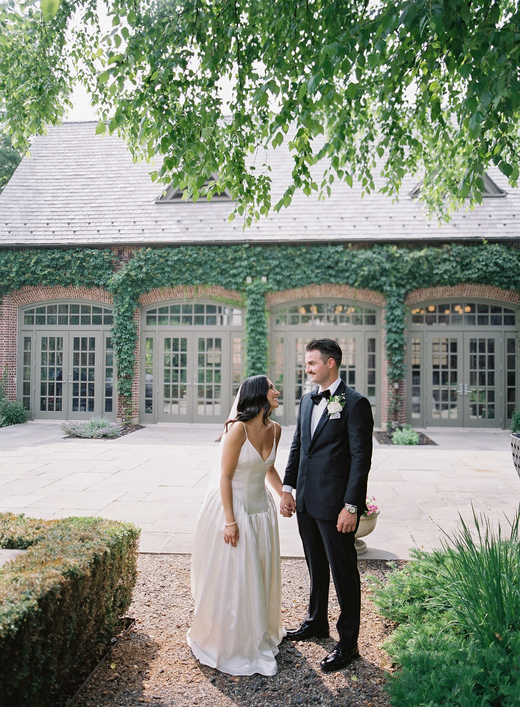 A bride and groom holding hands, smiling at each other outdoors in front of a building with large windows and ivy-covered walls.