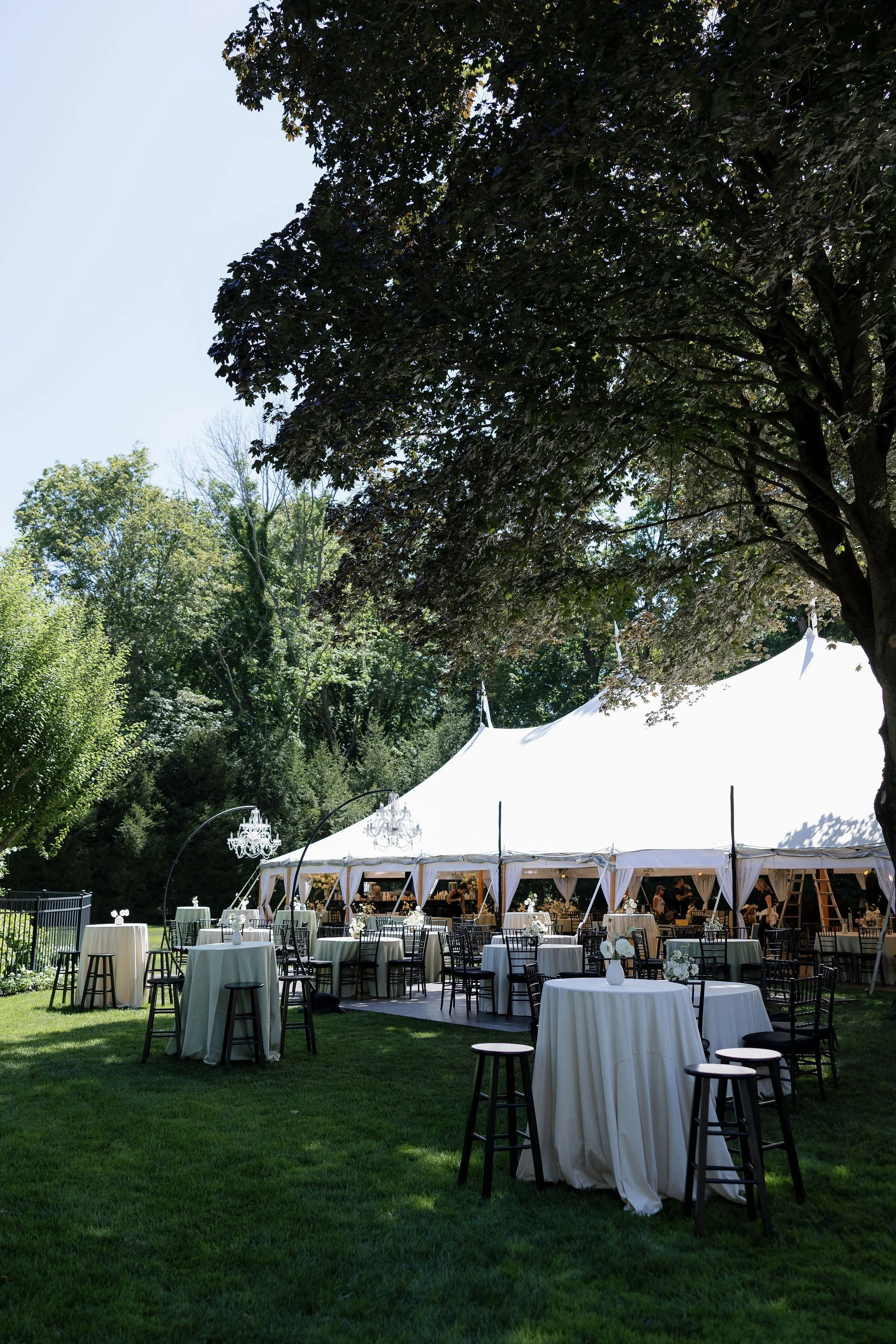Outdoor event setup with round tables covered in white tablecloths, black chairs, and a large white tent surrounded by trees and greenery.