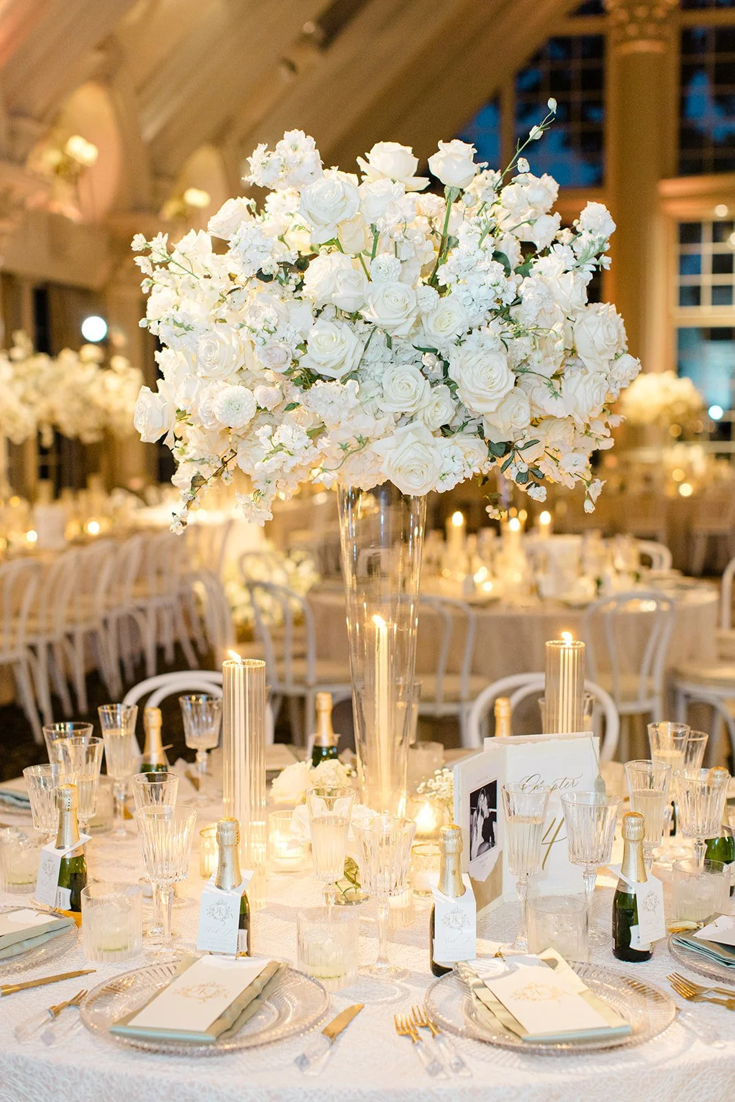 A table set for a wedding reception with a large centerpiece of white roses and flowers in a tall glass vase surrounded by candles, champagne bottles, and place settings with gold utensils and glassware, in a decorated banquet hall.