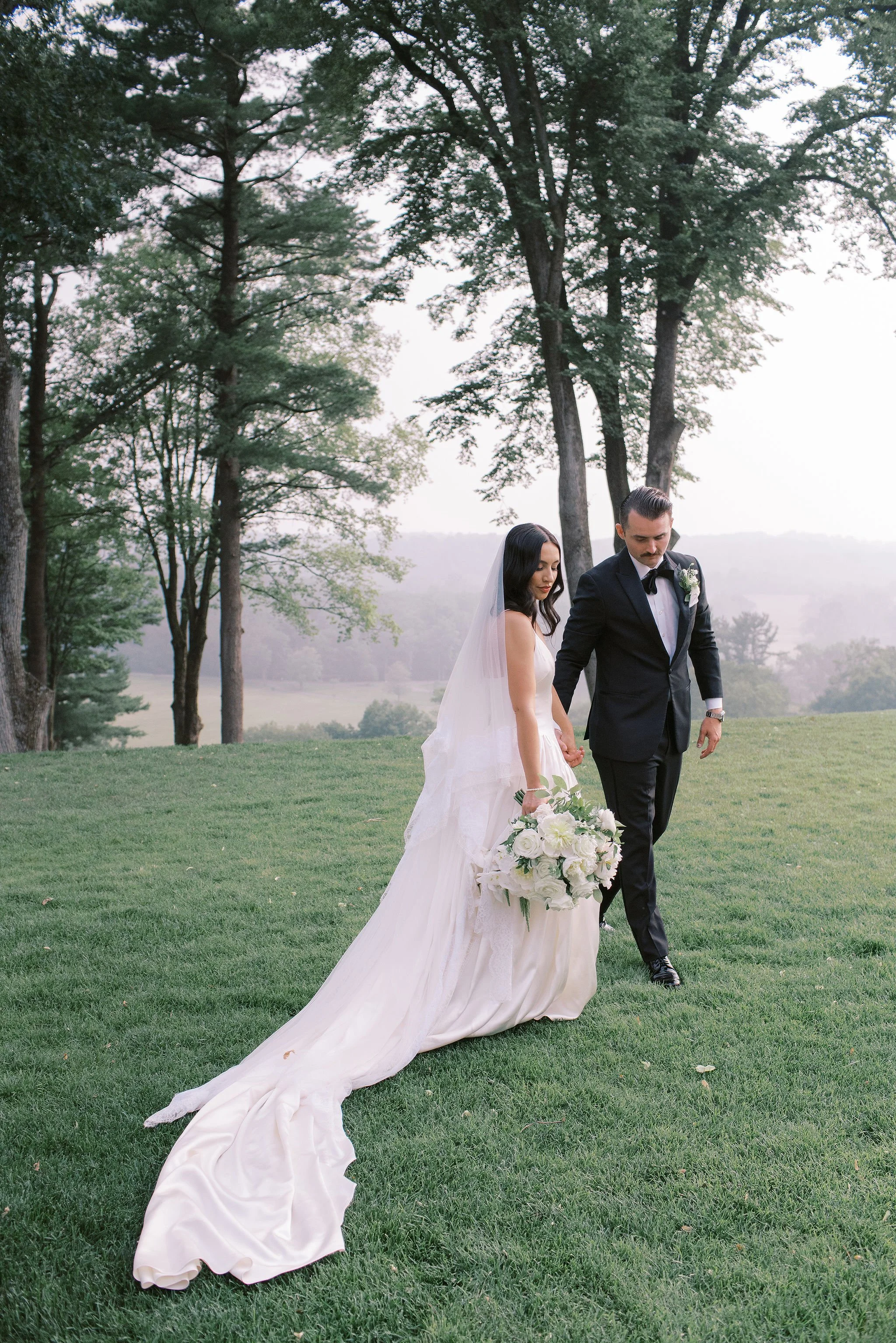 A bride and groom walking hand in hand on a grassy field with trees and a scenic landscape in the background, during a wedding photoshoot.