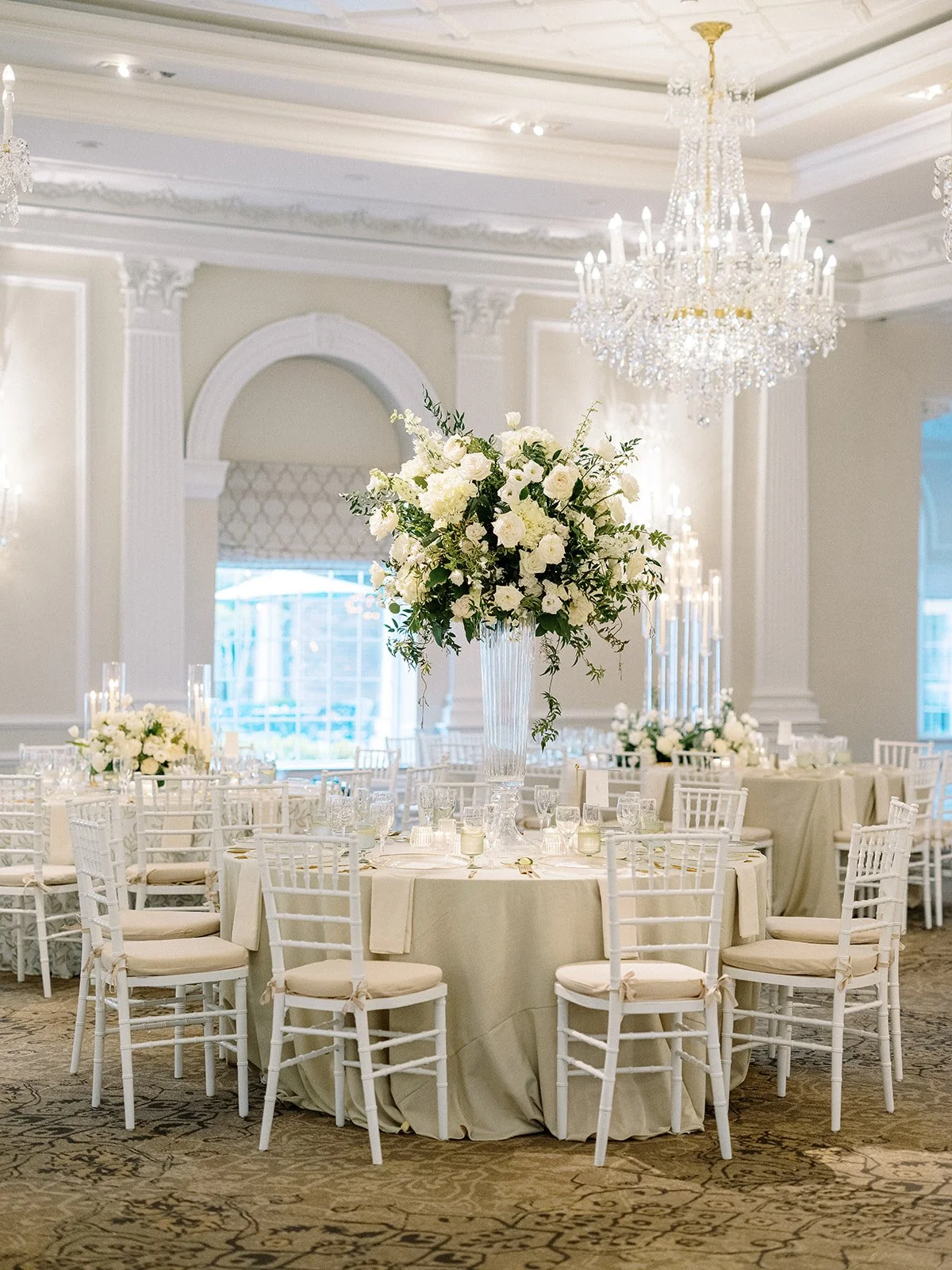 Elegant banquet hall with a tall floral centerpiece of white roses and greenery on a round table, surrounded by white chairs, crystal chandeliers, and soft lighting.