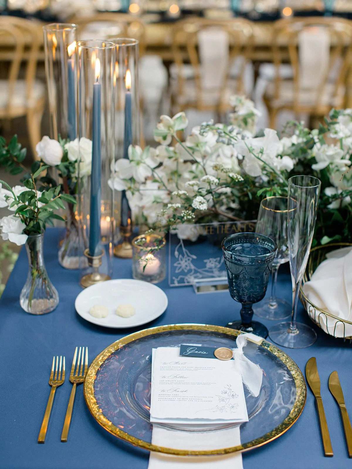 Elegant table setting with a blue tablecloth, gold flatware, a gold-rimmed glass plate with a menu, blue and clear glassware, white floral centerpiece, candles, and navy blue accents for a formal event.