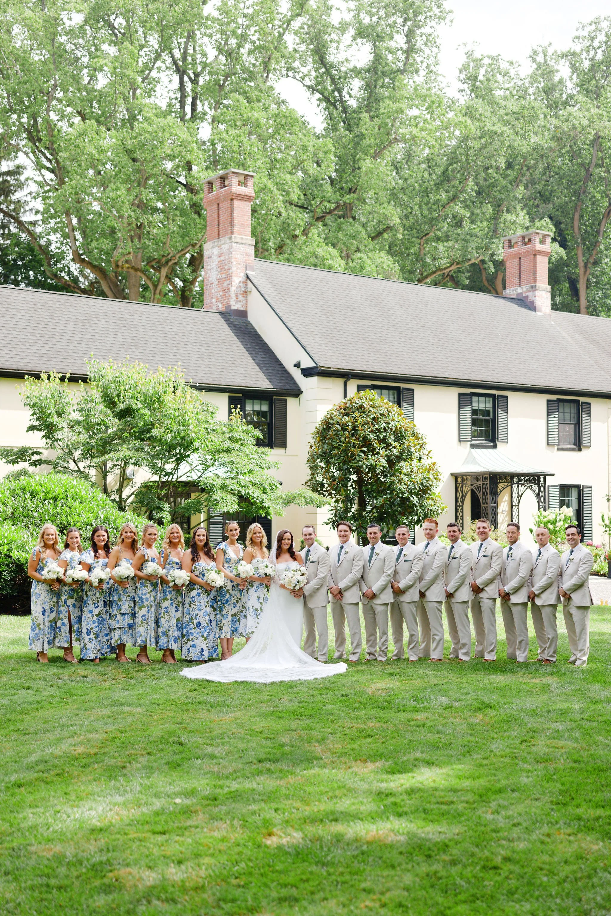 Wedding party photo with bridal couple in the center, bridesmaids in blue floral dresses on the left, groomsmen in beige suits on the right, standing on a well-manicured lawn in front of a large white house with black shutters, surrounded by greenery