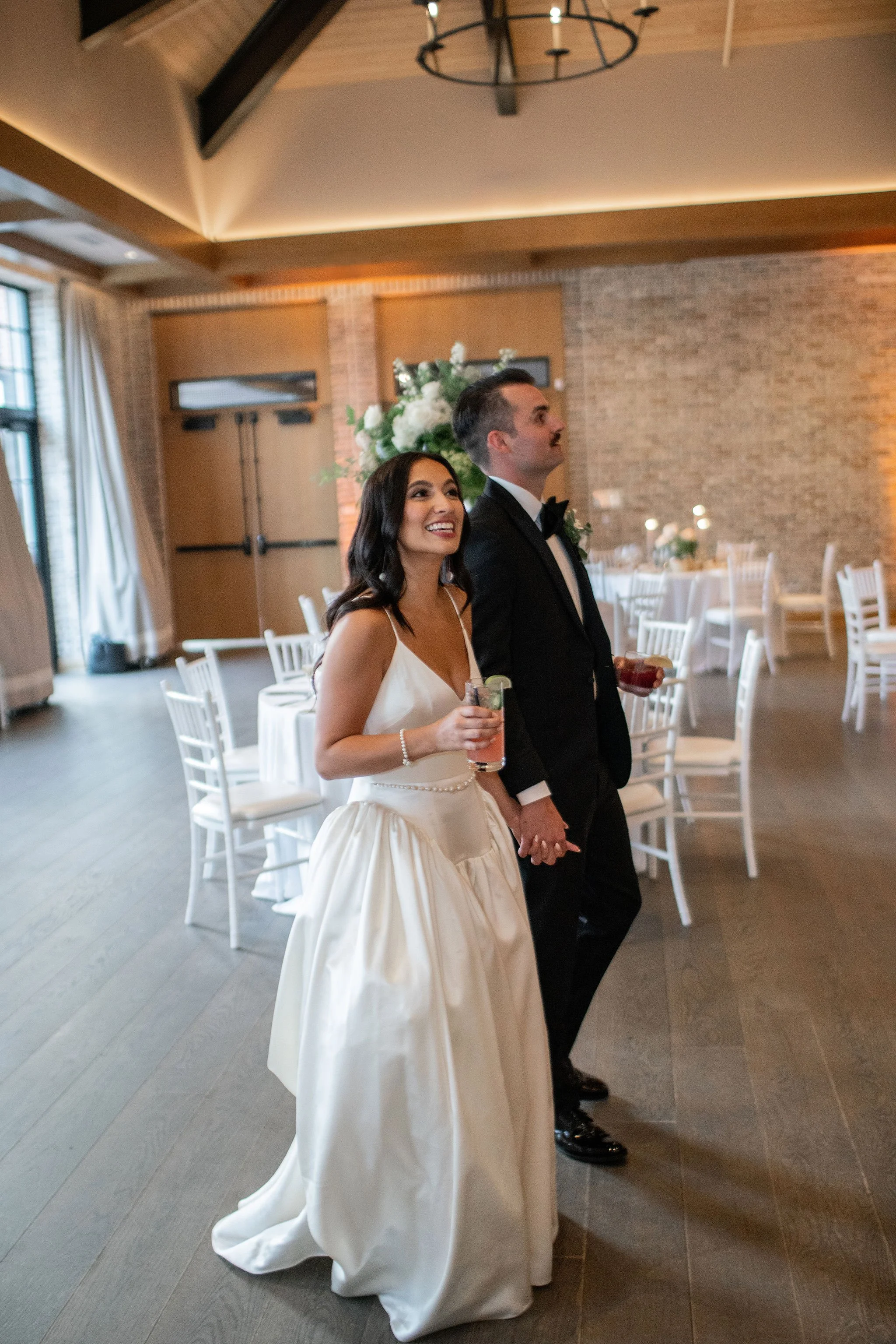 Bride and groom holding hands at their wedding reception, smiling, with white chairs and floral centerpieces in the background.