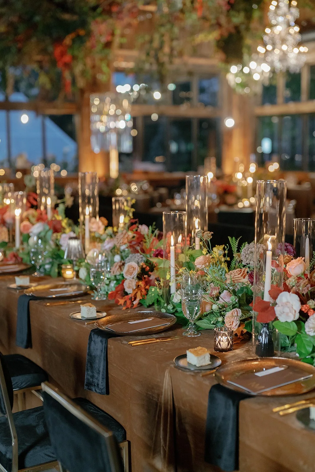 A beautifully decorated indoor reception table with floral arrangements, tall candles, and place settings with gold utensils and black napkins at a wedding or special event.