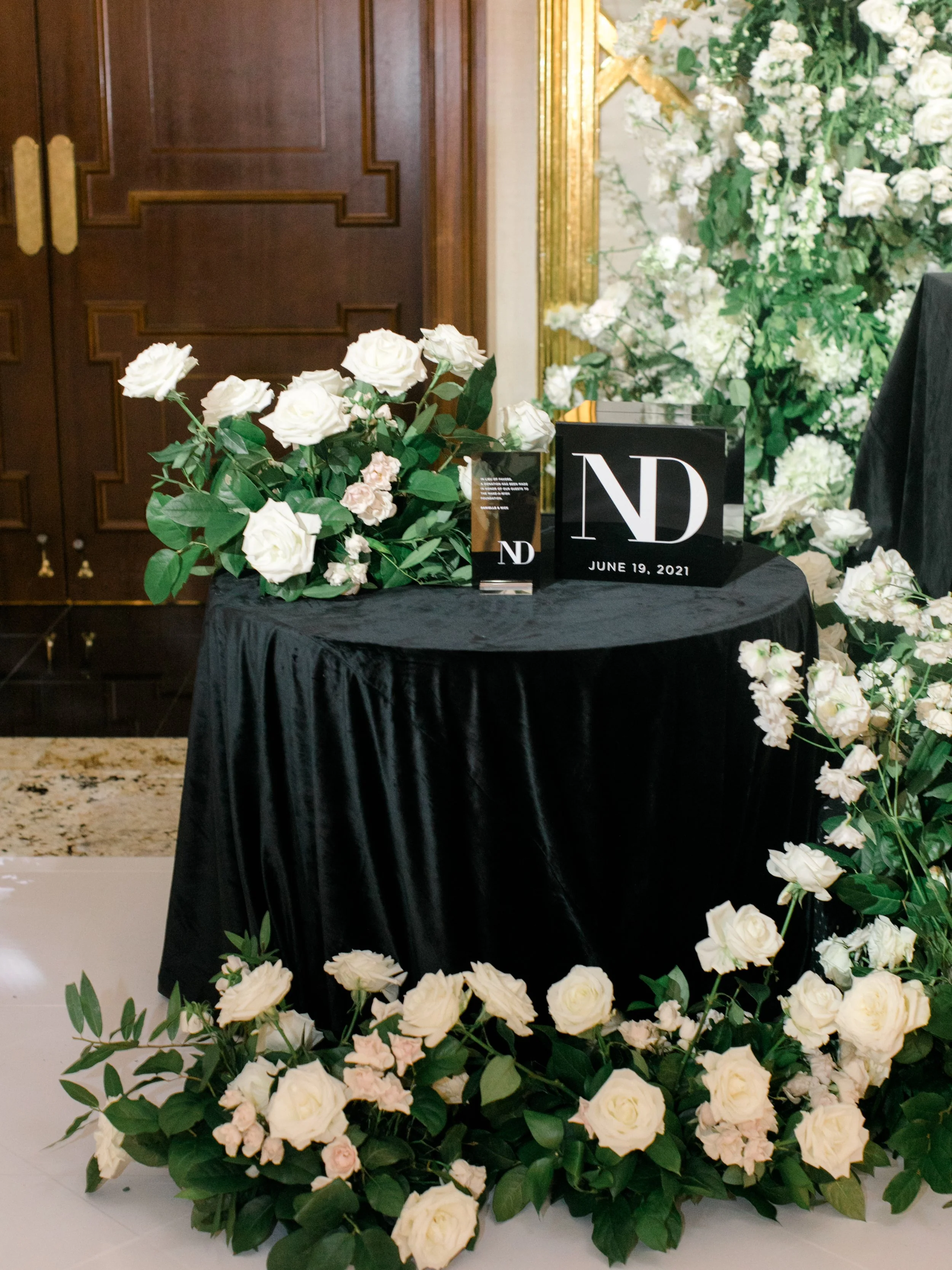 A black tablecloth-covered table decorated with white and light pink roses and greenery, with a black sign displaying the initials 'ND' and the date June 19, 2021, inside a room with wooden doors and cream-colored walls adorned with gold accents and 
