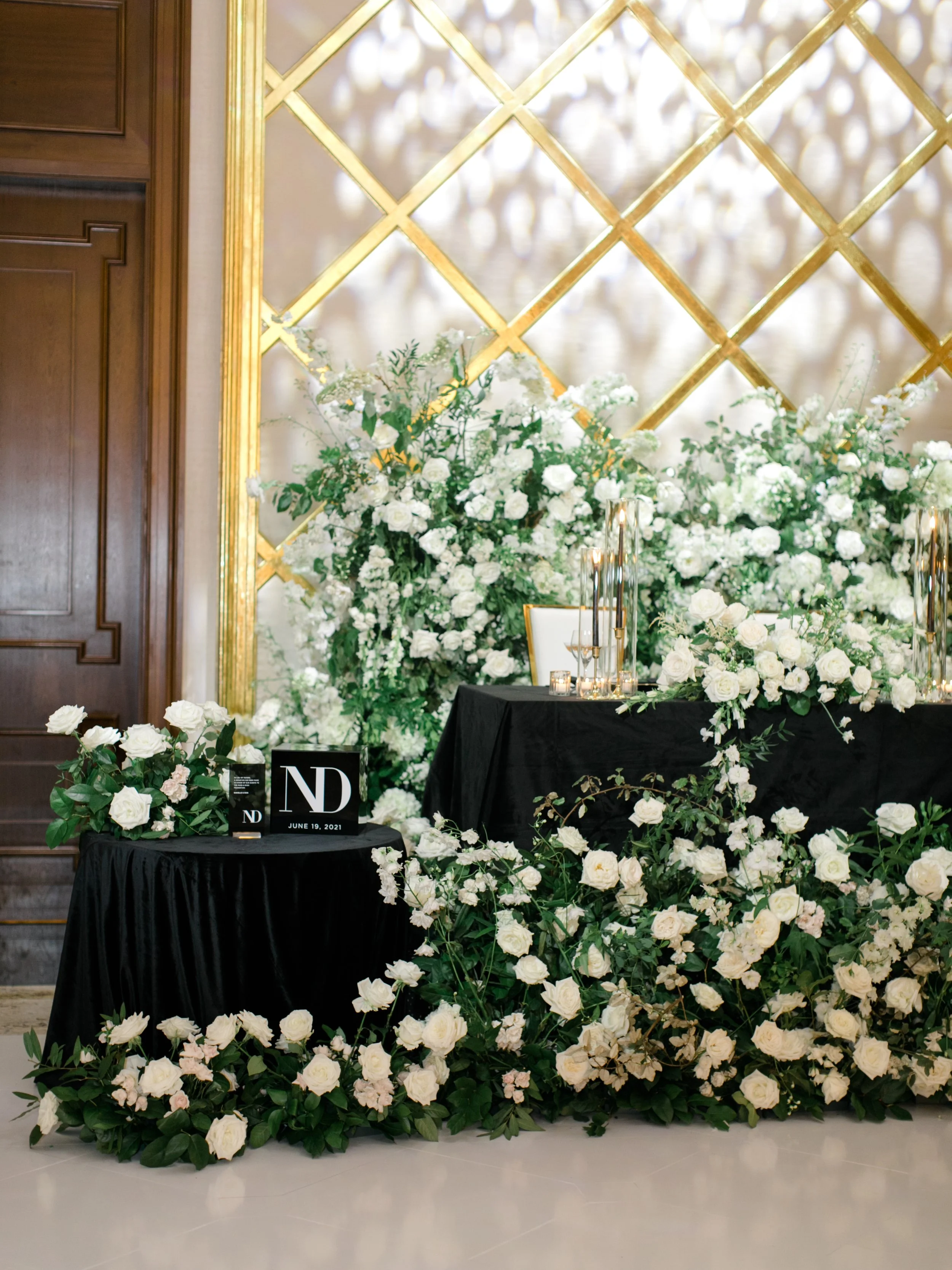 Elegant wedding or event reception table decorated with white flowers, greenery, and black tablecloths. Gold accents on the wall and flower arrangements create a luxurious atmosphere.
