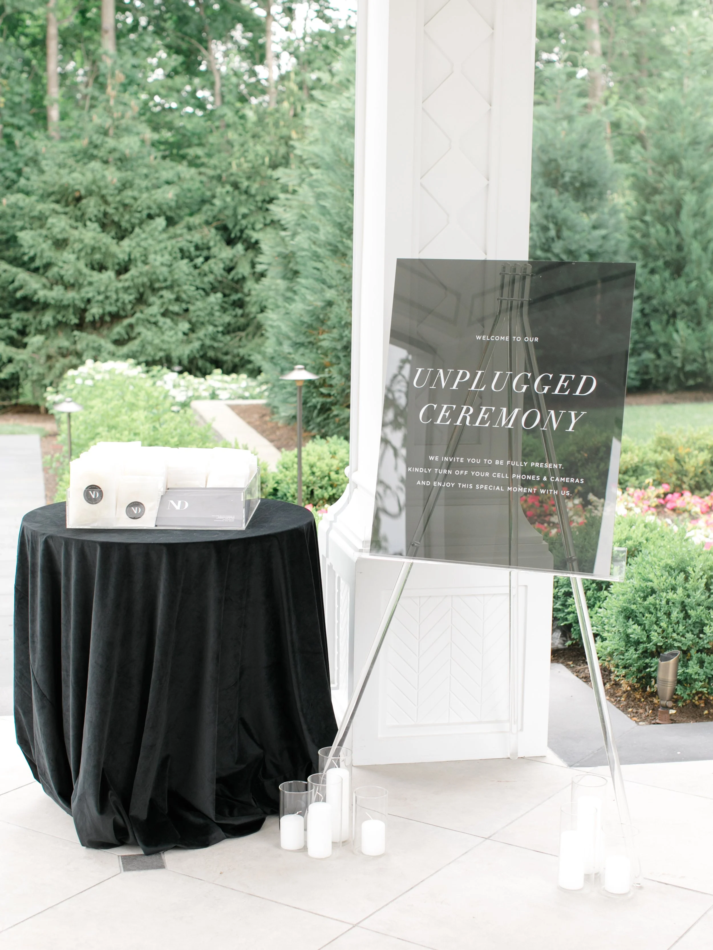 A sign on an easel at an outdoor wedding ceremony reads "Unplugged Ceremony" inviting guests to turn off cell phones and cameras. There is a round table covered with a black cloth with white candles and small jars labeled with initials next to a clea