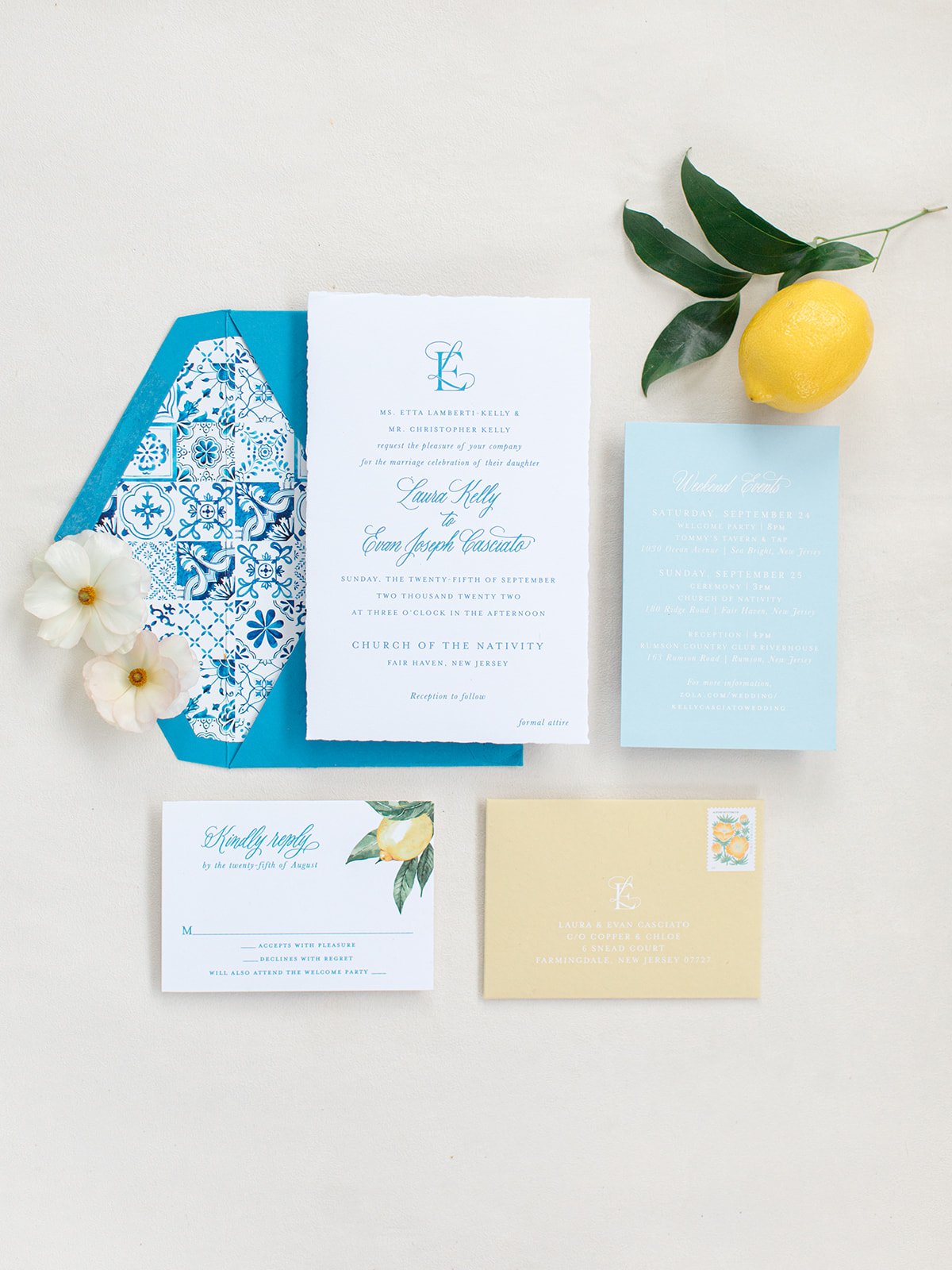 Wedding invitations with blue, white, and yellow accents, a lemon with leaves, white flowers, and a yellow envelope with a lemon stamp on a white surface.