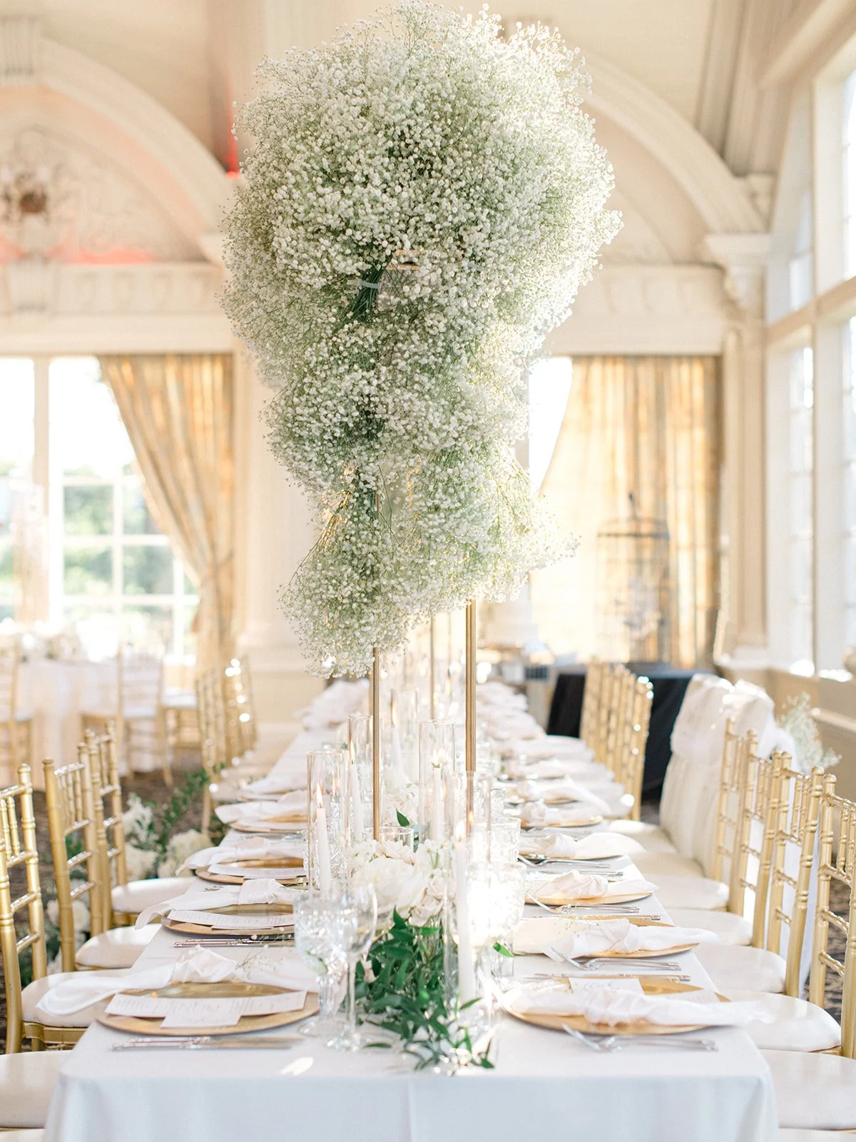 Elegant wedding table with gold Chiavari chairs, tall floral centerpieces of white baby's breath, surrounded by glass candleholders, set with white napkins, plates, and glassware in a bright, airy room with large windows and gold curtains.