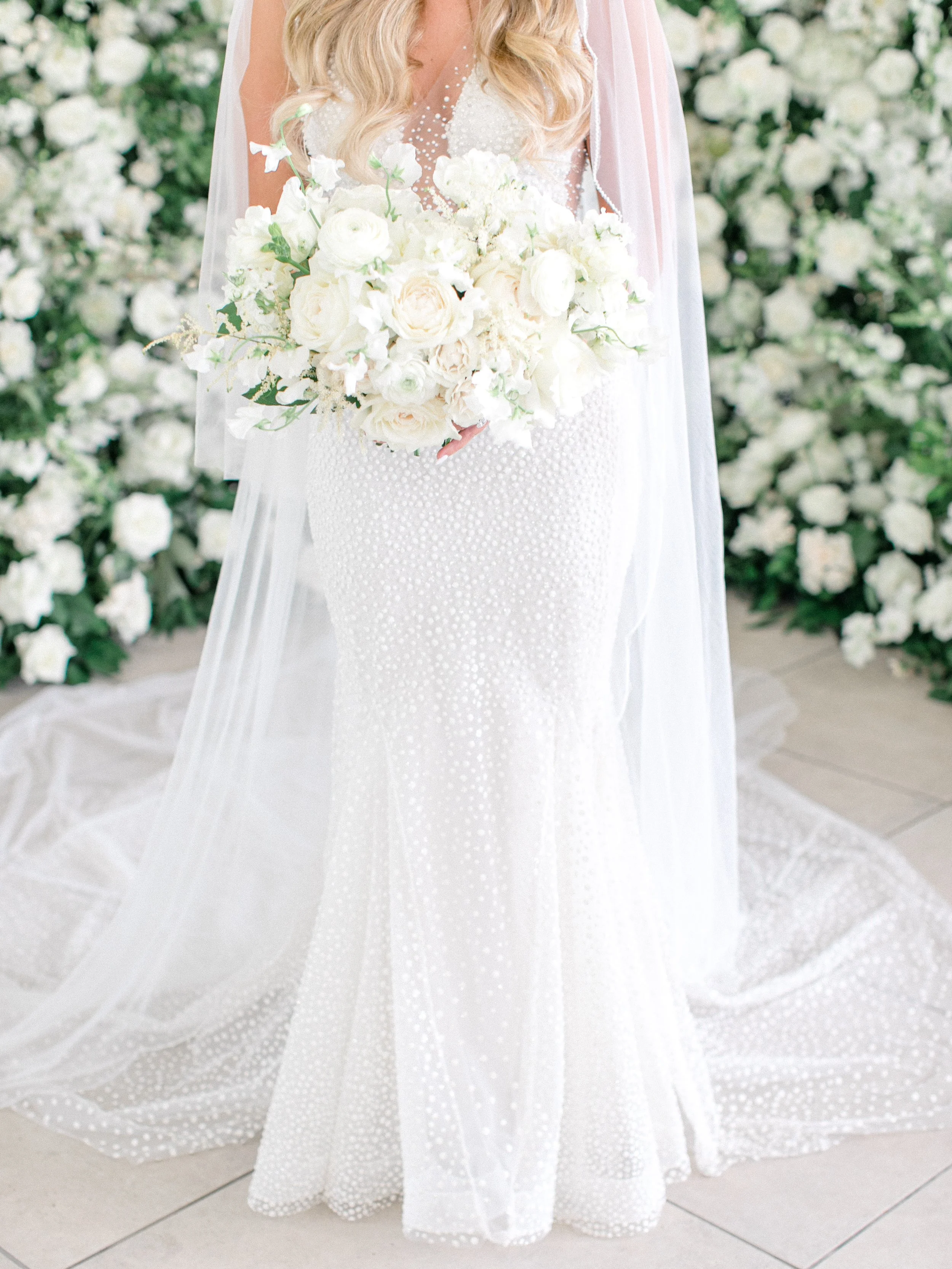 A bride in a fitted white wedding dress holding a bouquet of white flowers, standing in front of a wall of white flowers.