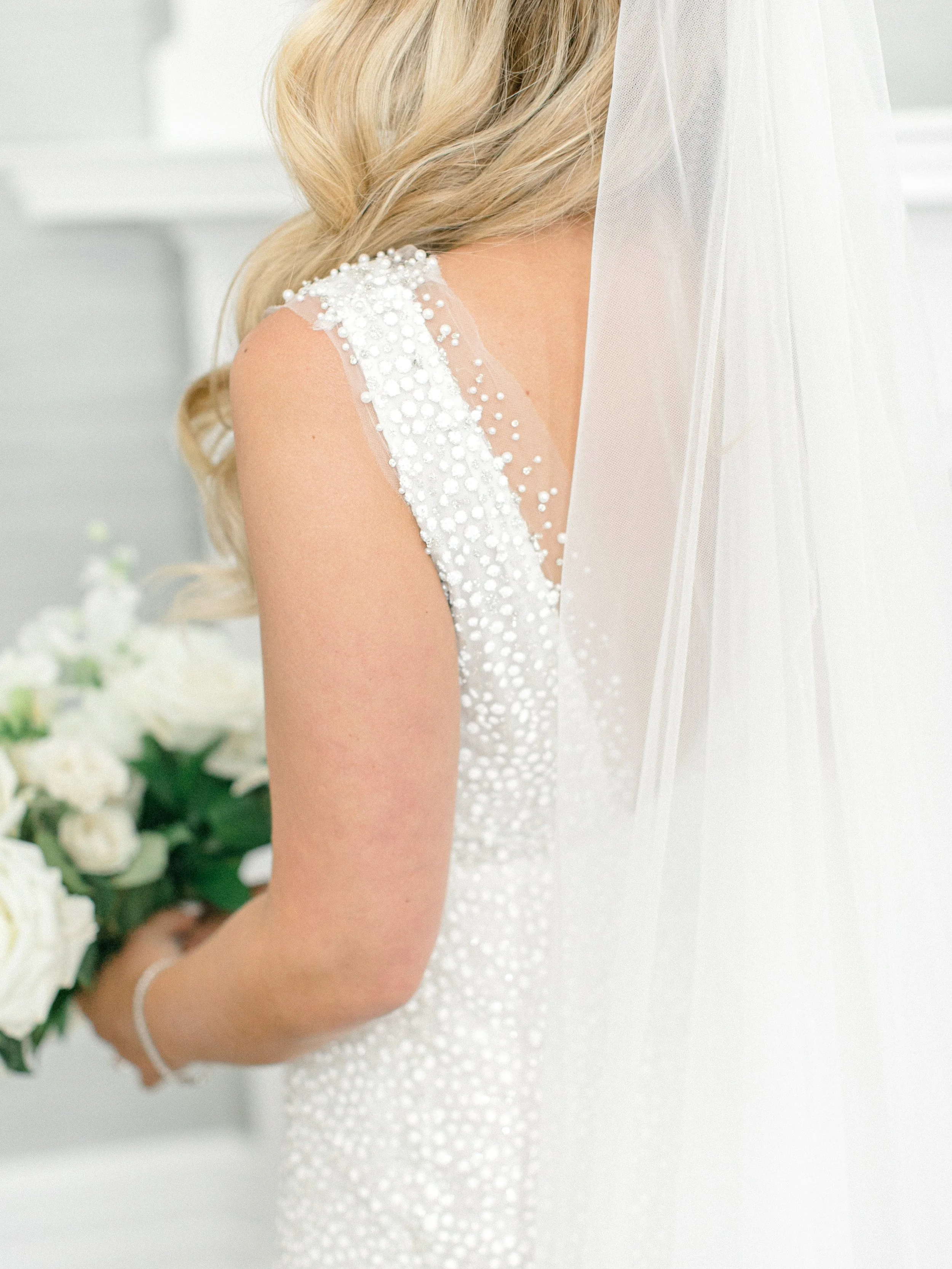 Close-up of a bride wearing a white wedding dress with pearl embellishments and a veil, holding a bouquet of white roses.