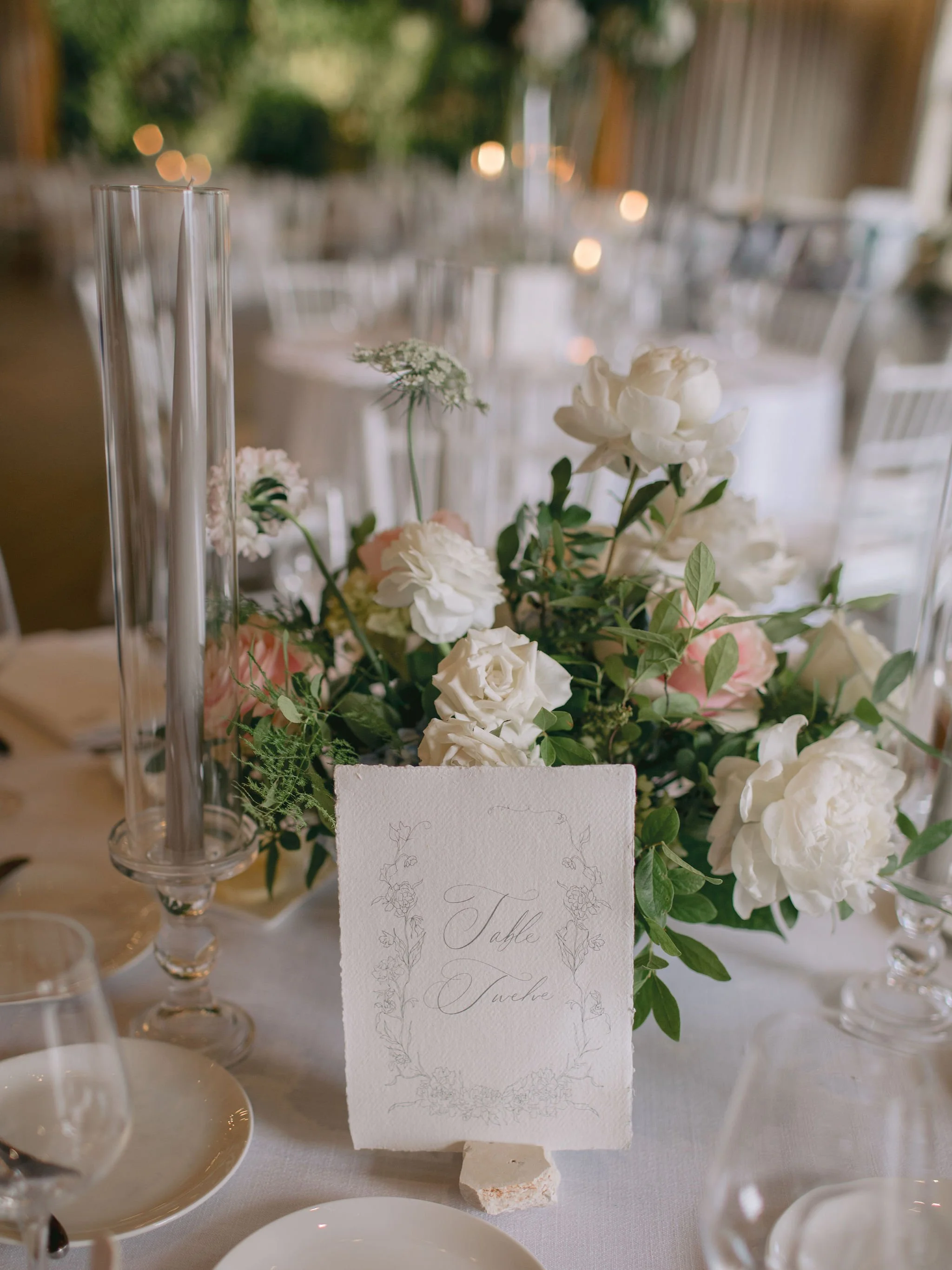 Elegant wedding table centerpiece with white roses, peonies, and greenery, with a table number card reading 'Table Twelve' in cursive, surrounded by glassware and candles.