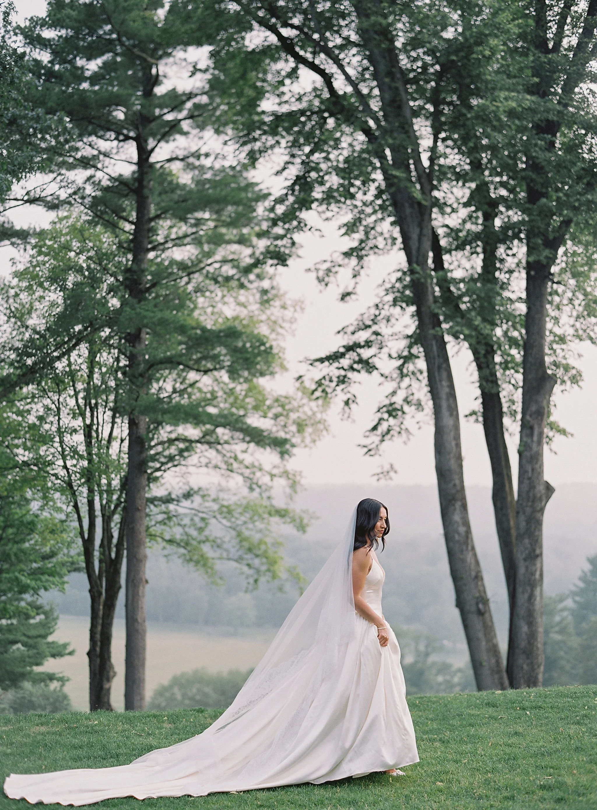 A bride in a white wedding gown with a long train and veil standing outdoors on green grass, surrounded by tall trees with a scenic backdrop of a misty landscape.