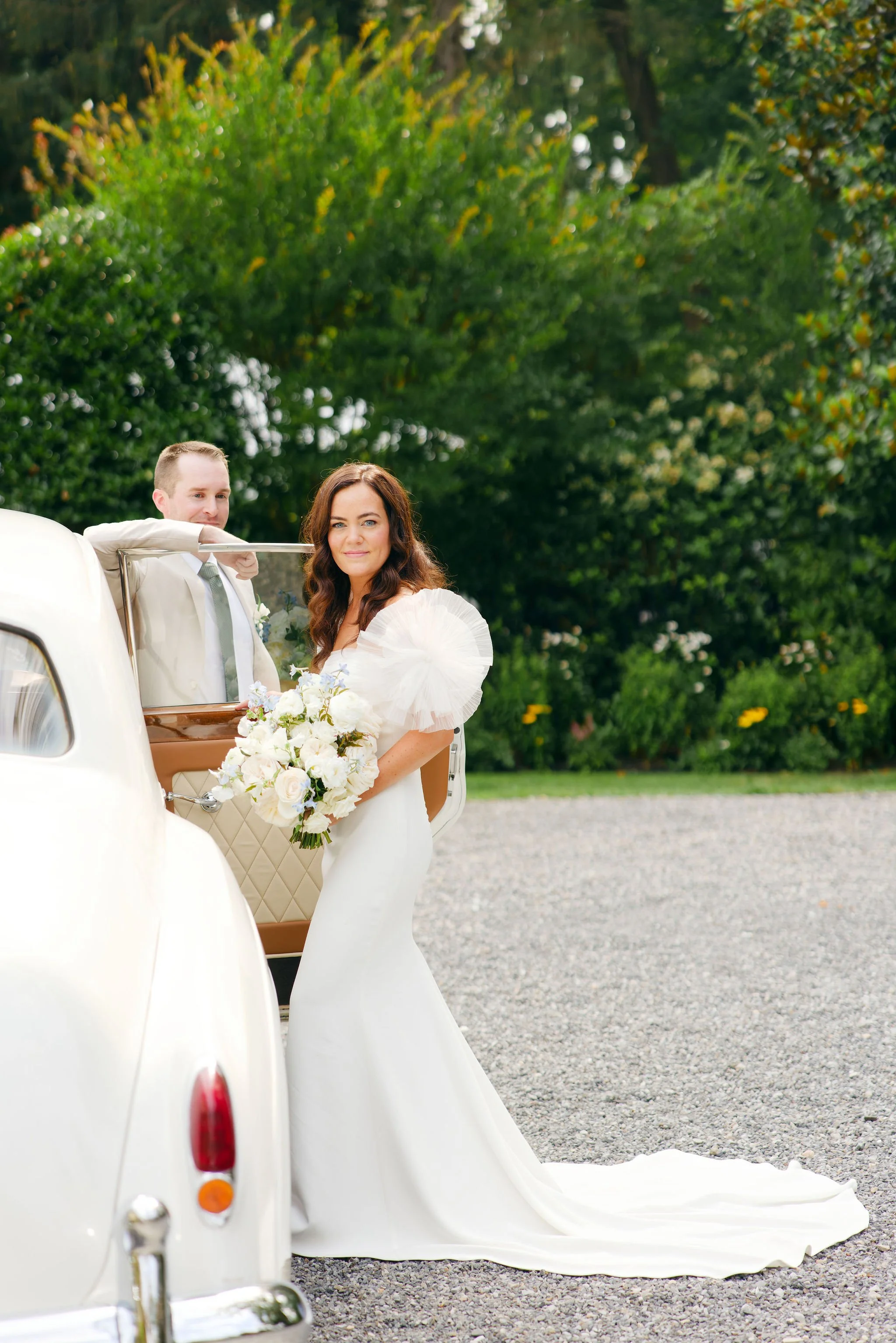 A bride in a white wedding gown holding a bouquet of white flowers and a groom in a suit standing beside a vintage white car outdoors with greenery in the background.