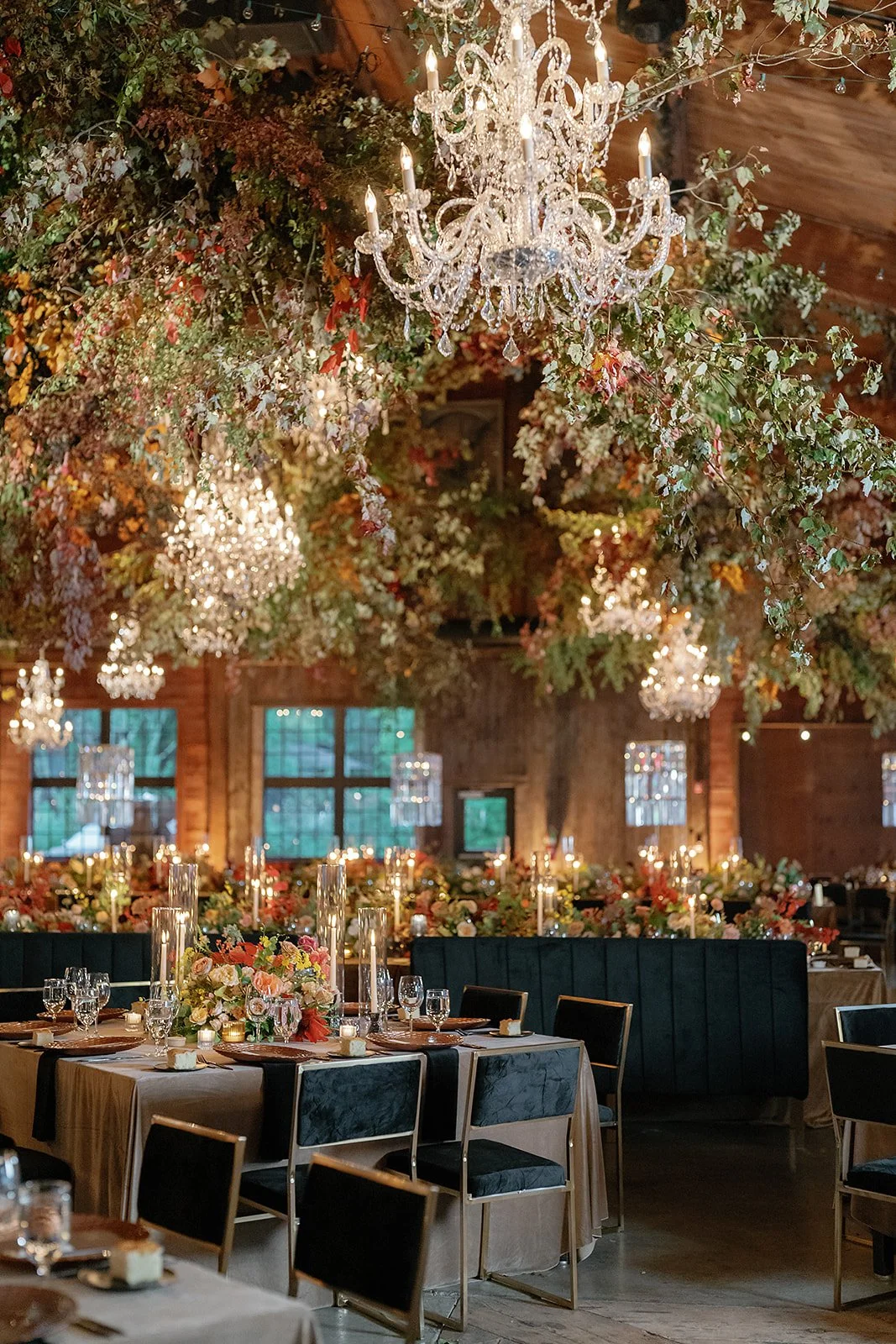 Elegant wedding reception in a rustic wooden barn with floral ceiling decorations, chandeliers, and arranged dining tables with floral centerpieces and candles.