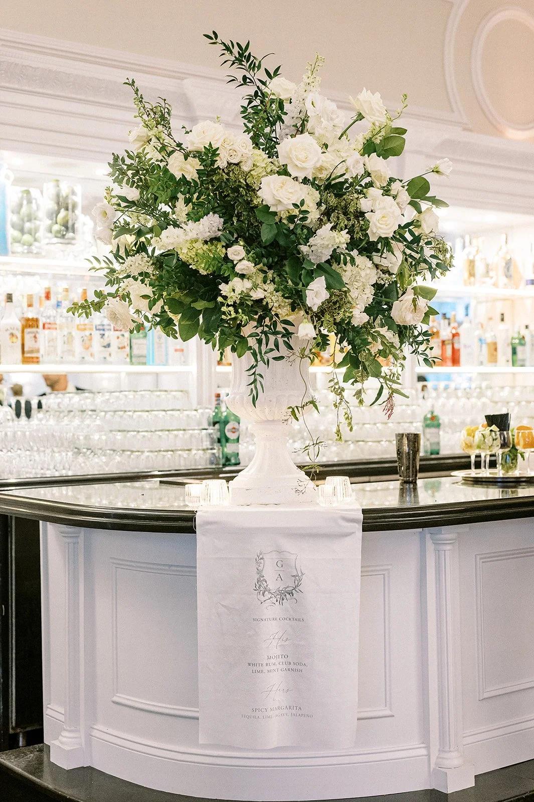 A large floral arrangement with white flowers and green foliage on a white bar counter, with a cocktail menu hanging on the front of the counter.