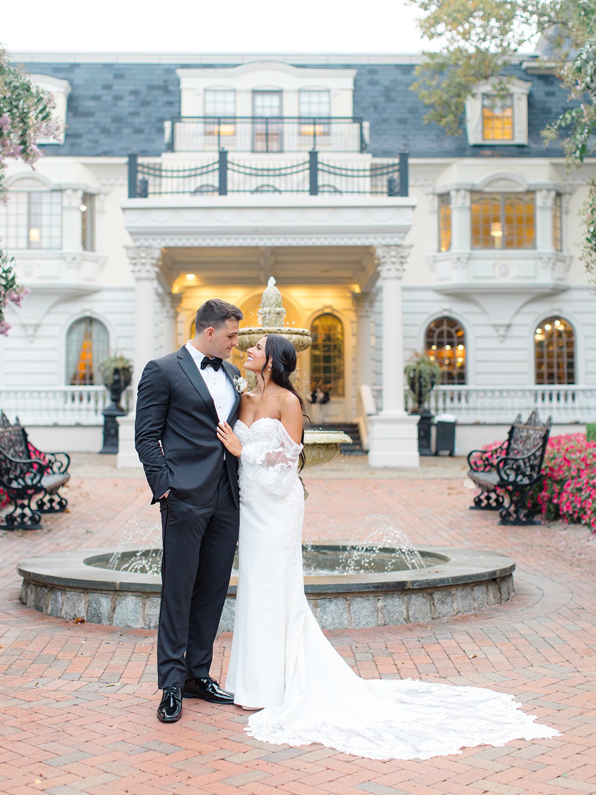 A bride and groom stand close together in front of a fountain, smiling at each other, in front of a large, white, historic mansion with ivy and pink flowers nearby.
