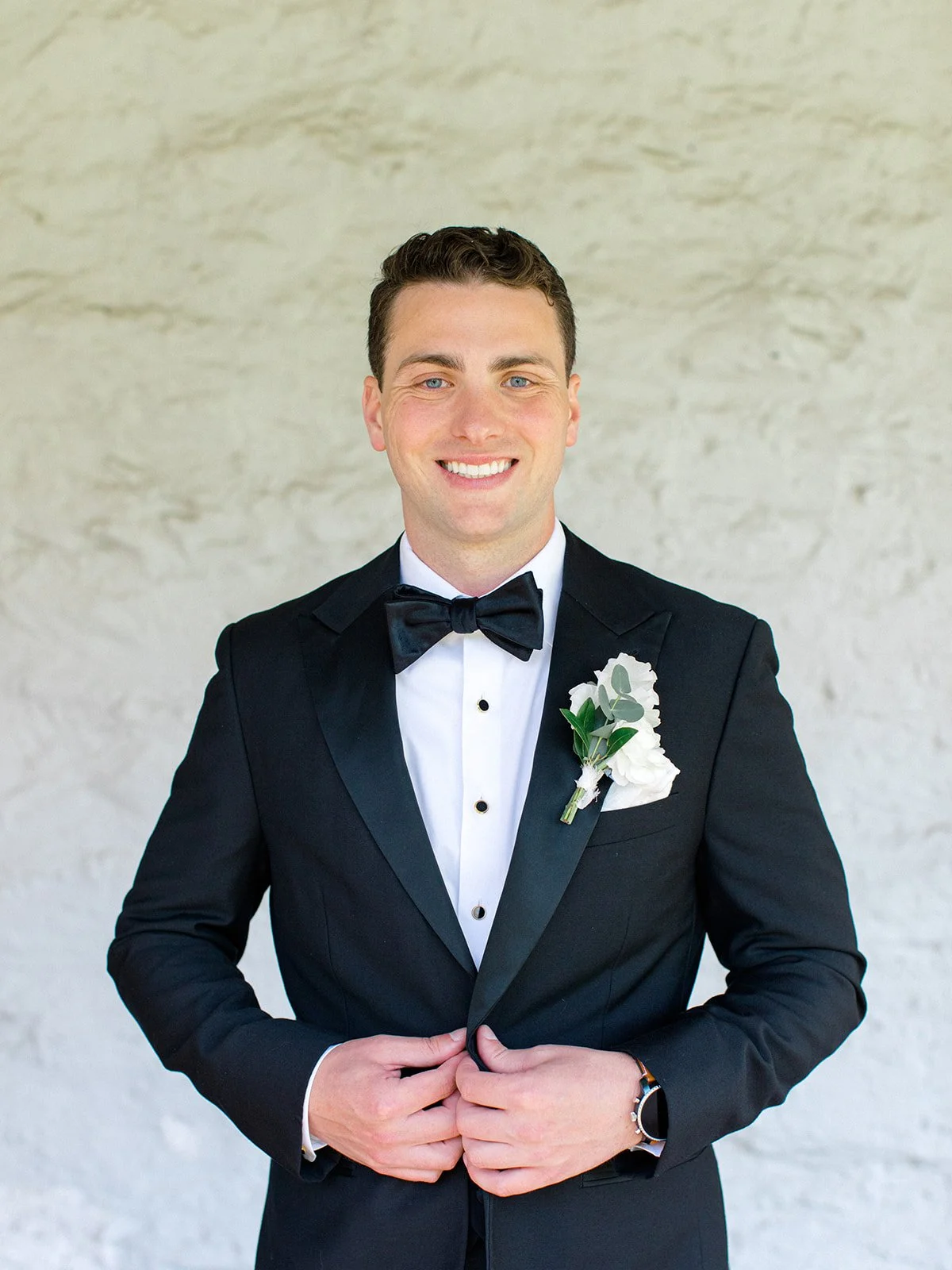 A groom in a black tuxedo with a white shirt and black bow tie, smiling, standing against a light-colored textured wall.