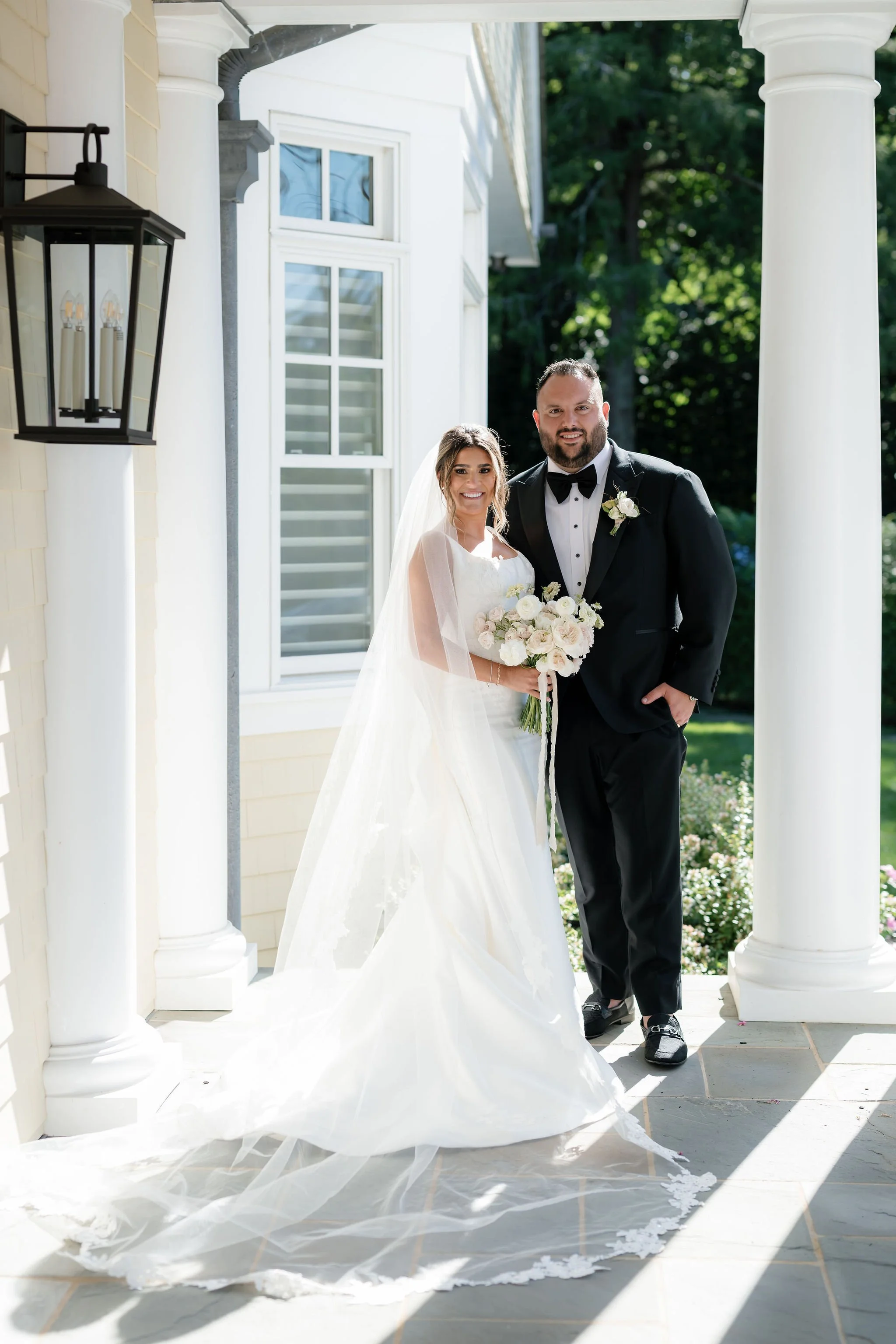 A bride and groom stand together outdoors on a sunny day, dressed in wedding attire. The bride holds a bouquet of white and pink flowers, and both are smiling at the camera. The setting features white columns and a house in the background.