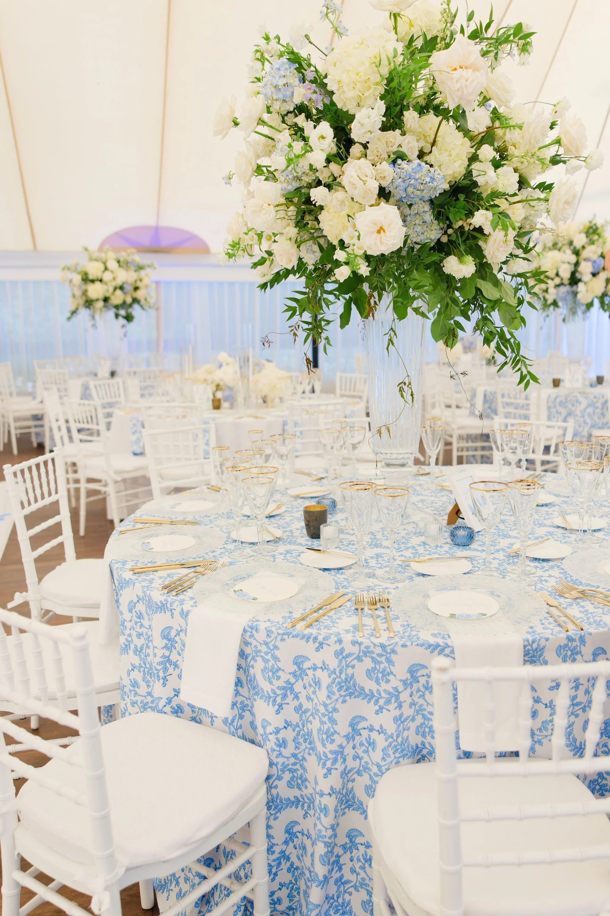 Elegant banquet table decorated with a large floral centerpiece featuring white and light blue flowers, set with white plates, gold utensils, and glasses with gold rims, in a bright venue with multiple similar tables and white chairs.
