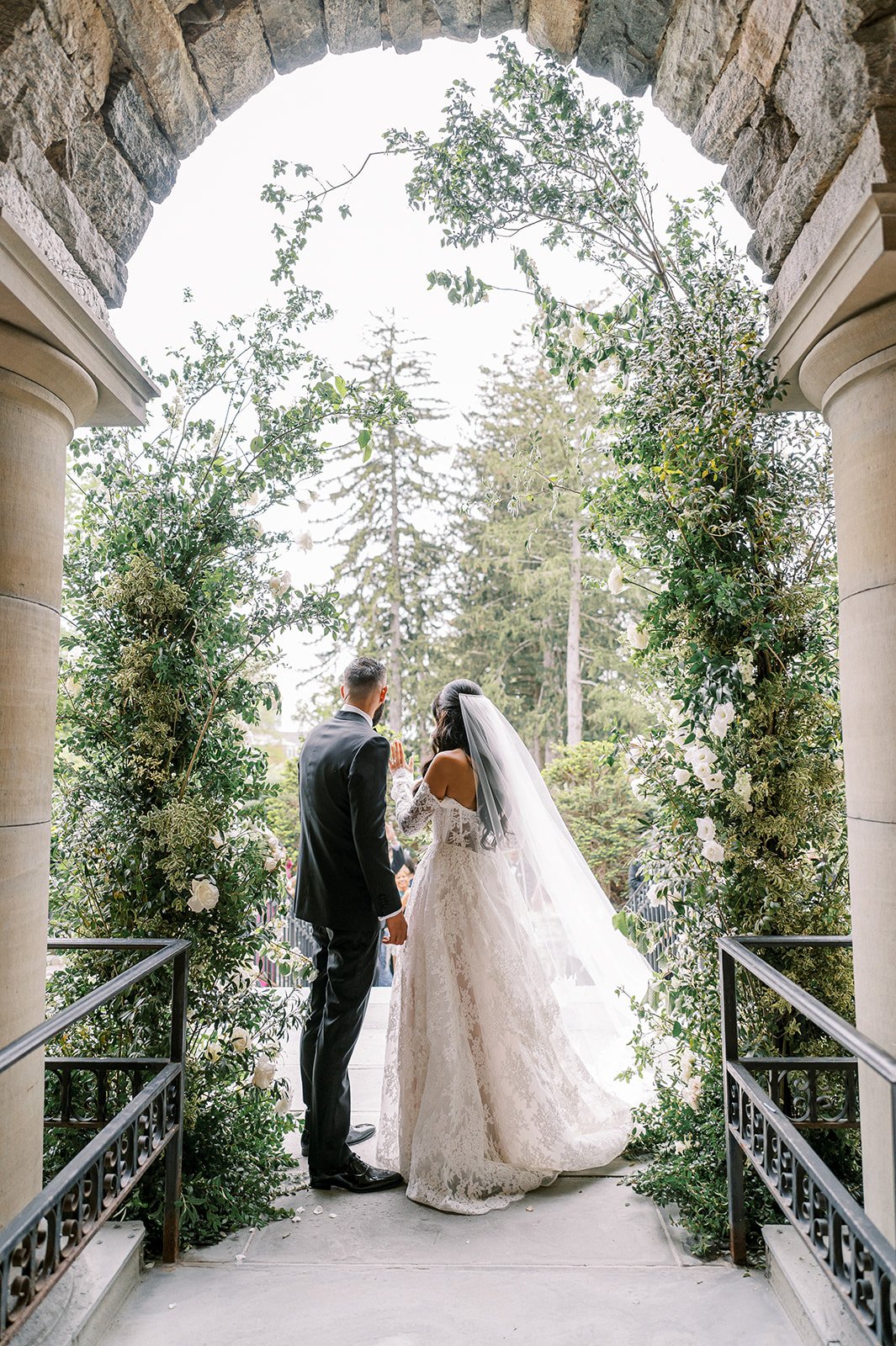 Bride and groom exchanging vows under a stone archway decorated with greenery and white flowers, with trees in the background.
