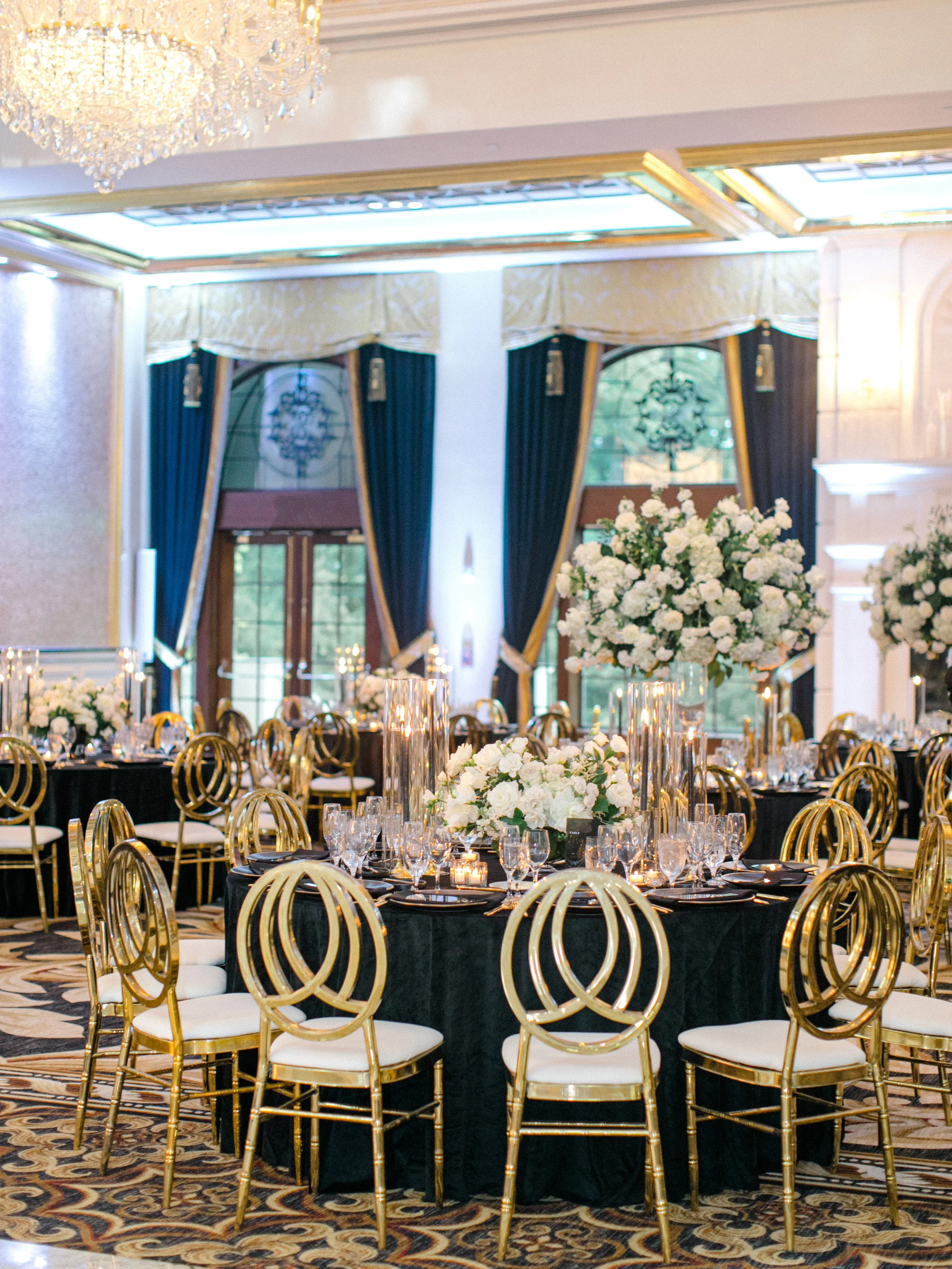 Elegant banquet hall decorated with black tablecloths, tall glass candles, and white floral centerpieces, with gold chairs and large windows with dark curtains.