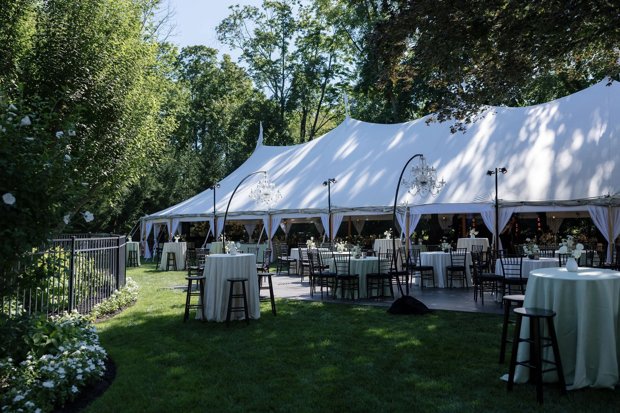 An outdoor wedding reception area under a large white tent, with round tables draped in white tablecloths, black chairs, and floral centerpieces, surrounded by green trees and grass.