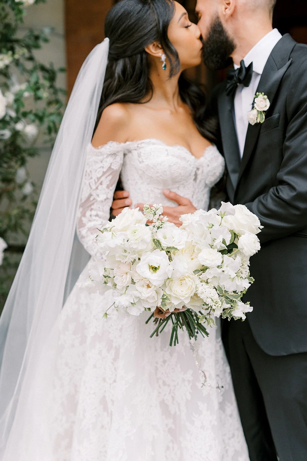 A bride and groom share a kiss at their wedding, with the bride holding a large bouquet of white flowers and wearing a lace off-shoulder wedding dress, while the groom is in a tuxedo with a boutonniere.