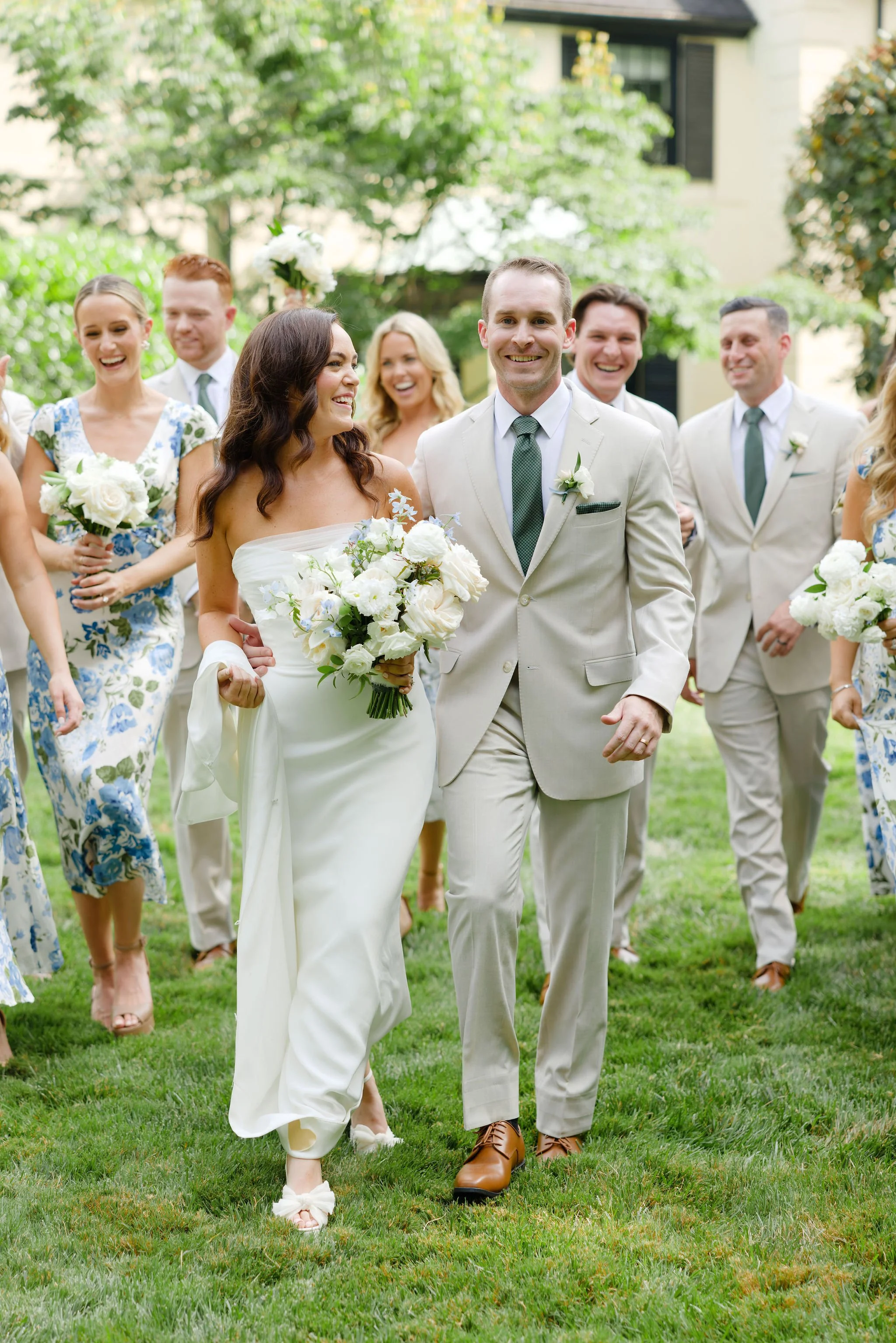 Wedding couple walking on grass with friends and family in the background, outdoors, greenery, smiling, bride holding bouquet, groom in beige suit.
