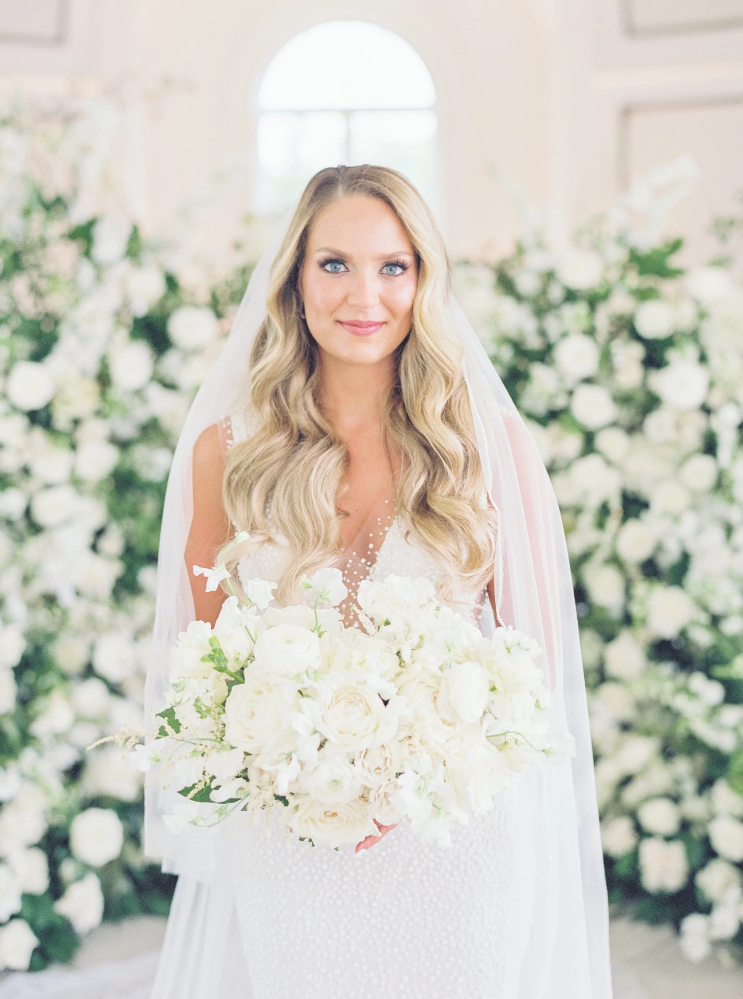 A bride with long wavy blonde hair and blue eyes wearing a white wedding dress and veil, holding a large bouquet of white flowers, standing in front of a background decorated with white flowers.